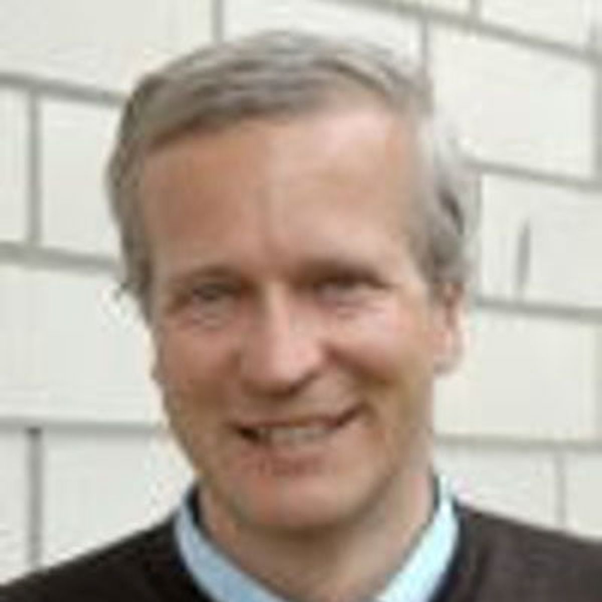 Middle-aged man with gray hair smiling at camera against white brick wall background.