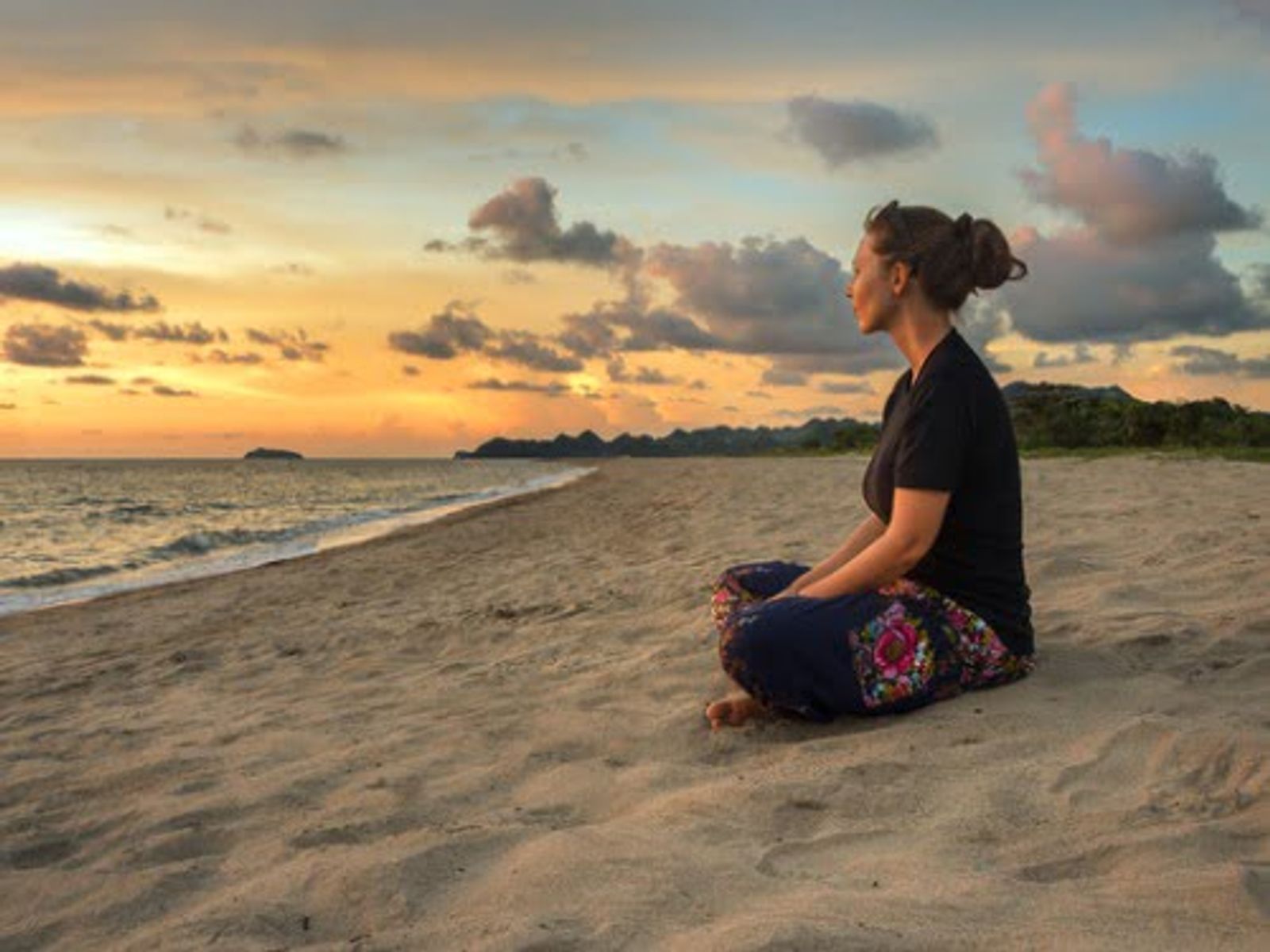 Woman in black shirt sitting on beach at sunset, facing ocean.
