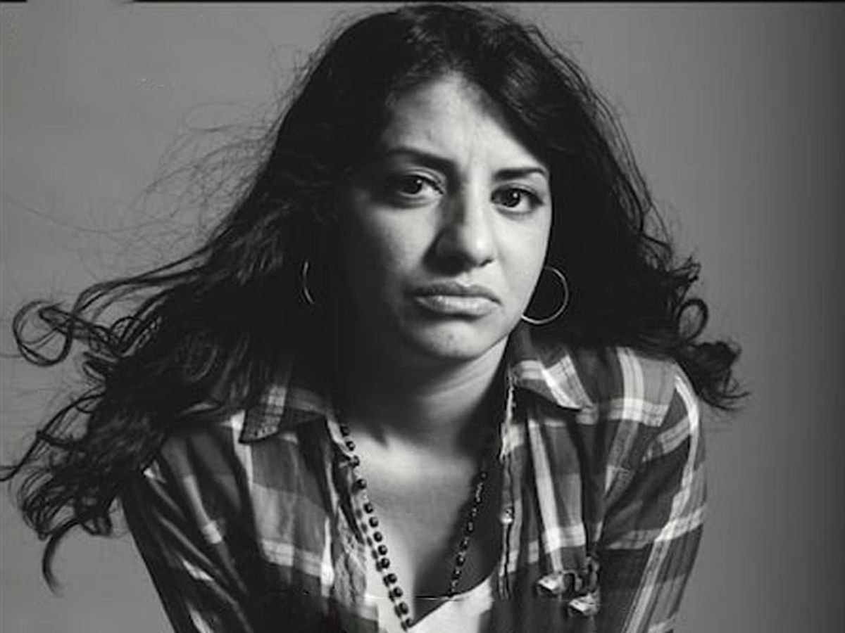 Black and white portrait of woman with long dark hair wearing plaid shirt and hoop earrings.