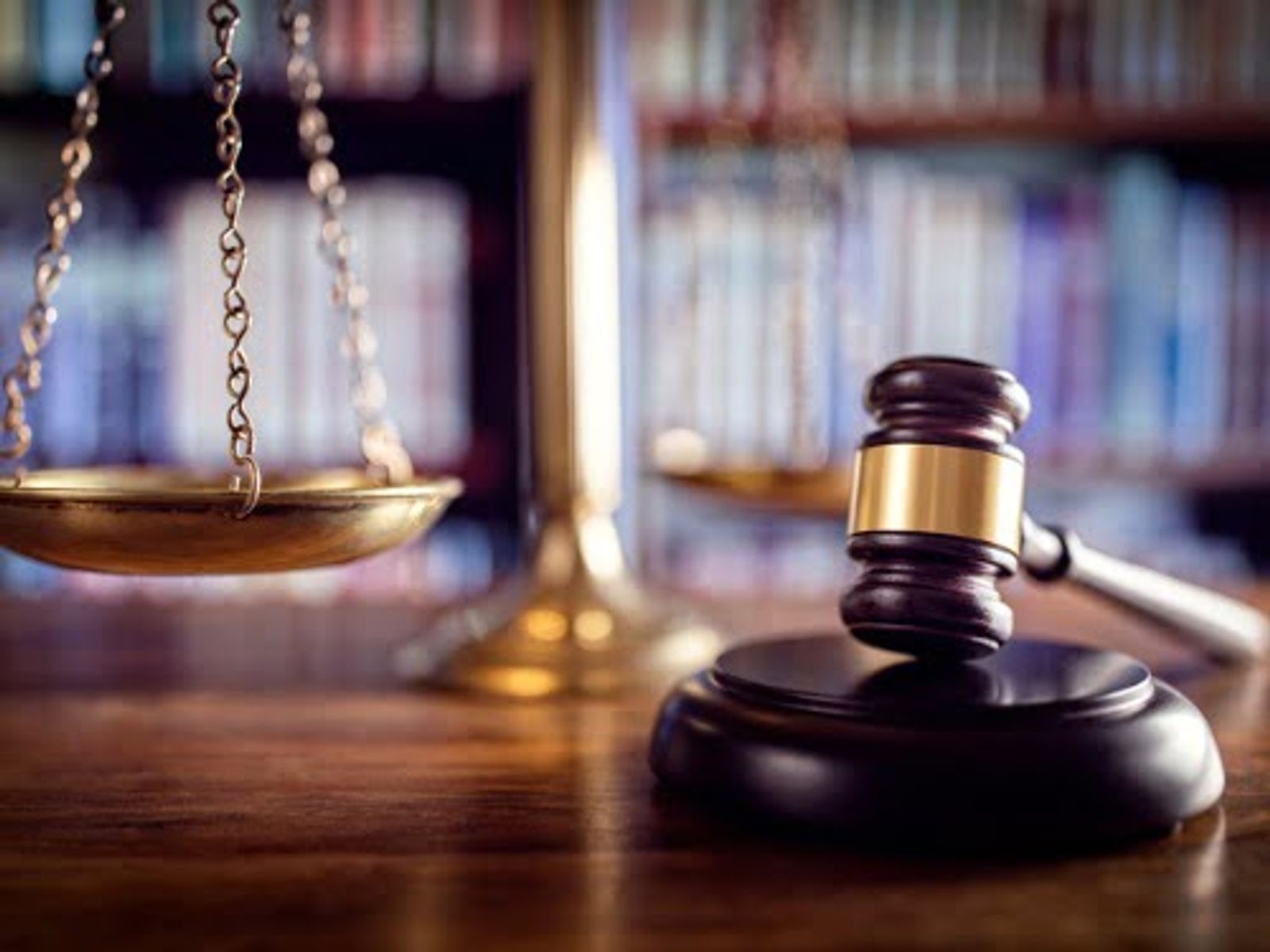 Mazo y balanza de la justicia sobre un escritorio de madera, con estanterías de una biblioteca jurídica desenfocadas al fondo.