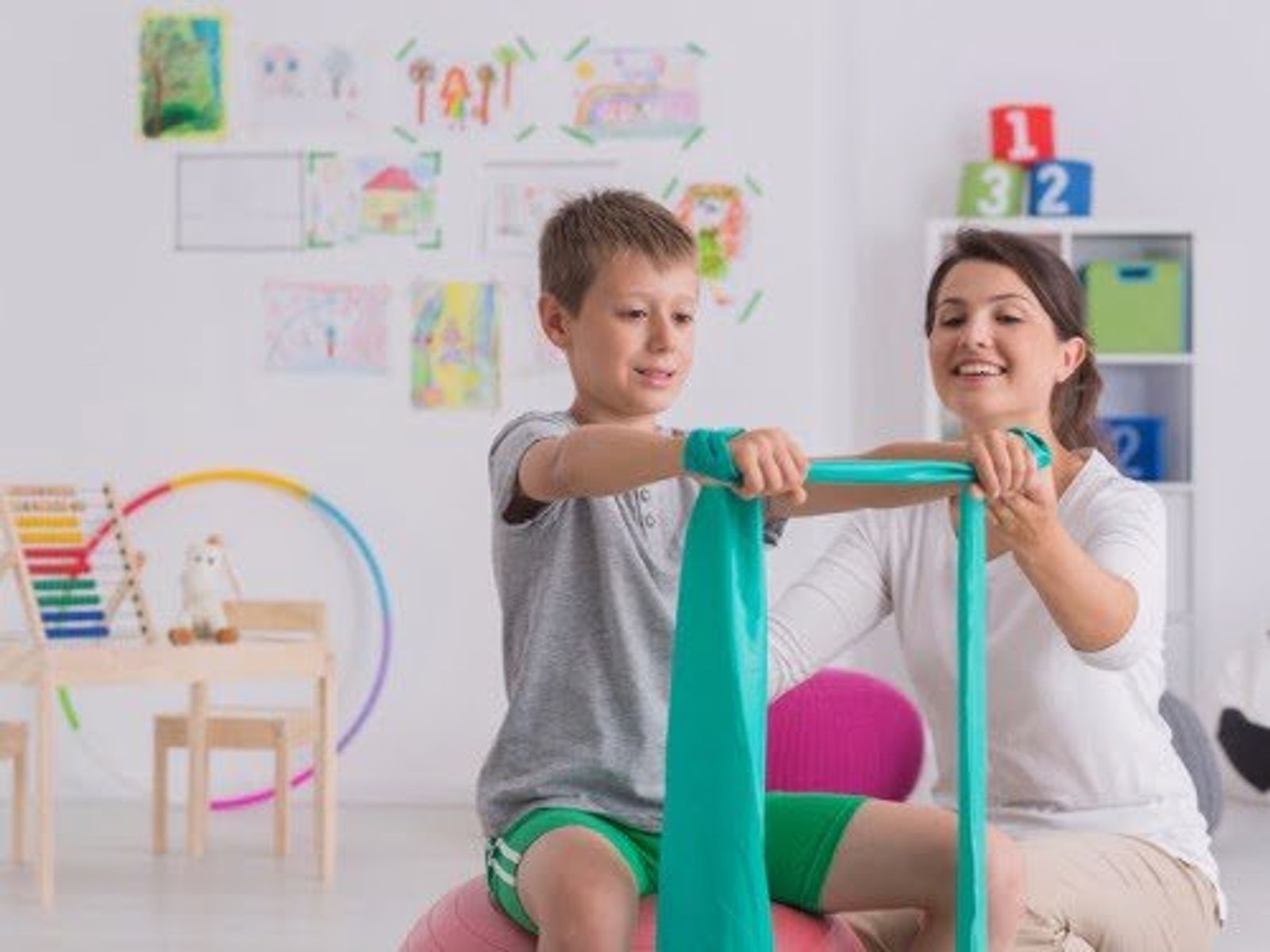 Woman and young boy using resistance band in colorful playroom with educational materials.