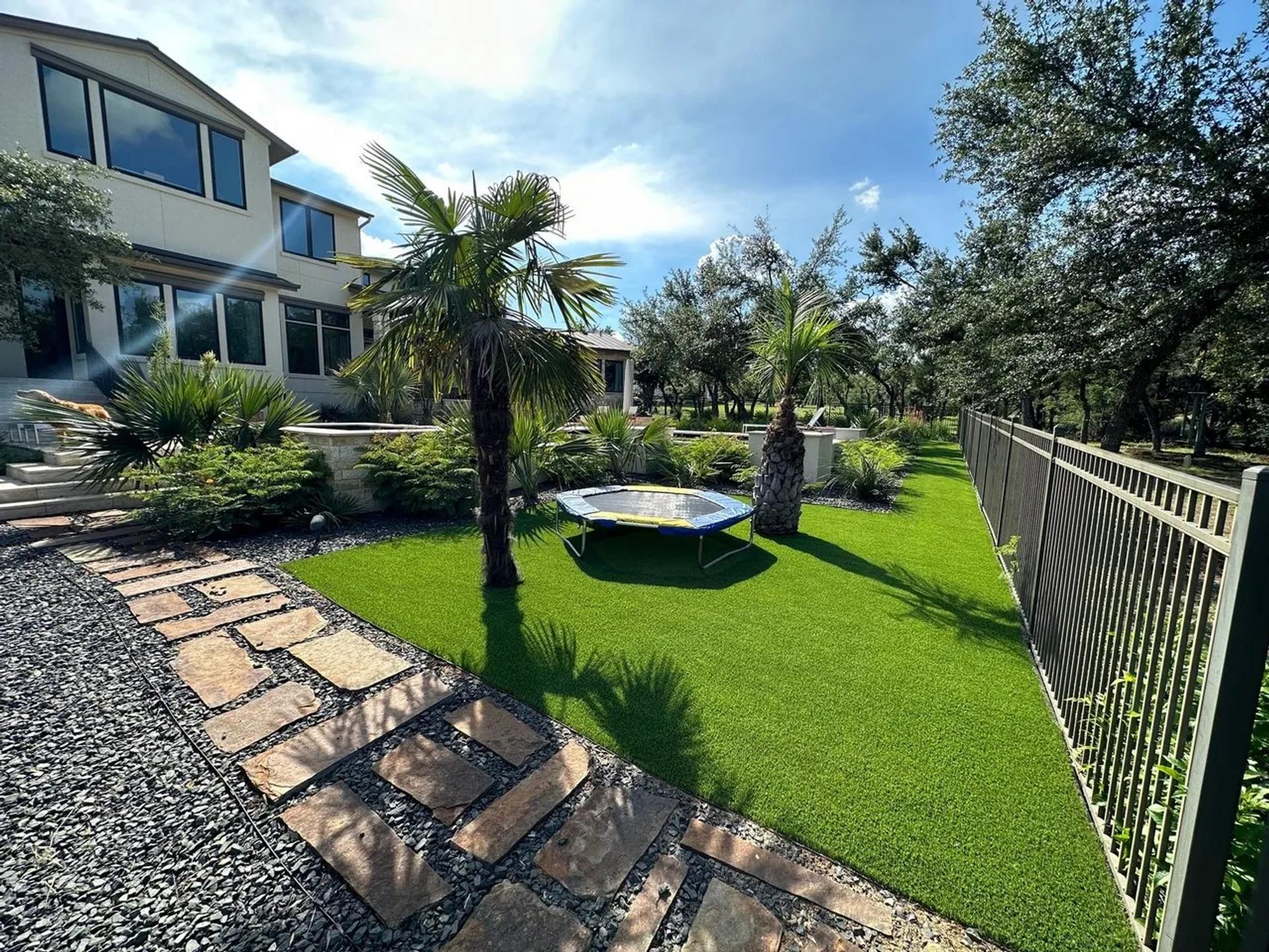 A trampoline surrounded by palm trees in artificial turf and a stone walkway