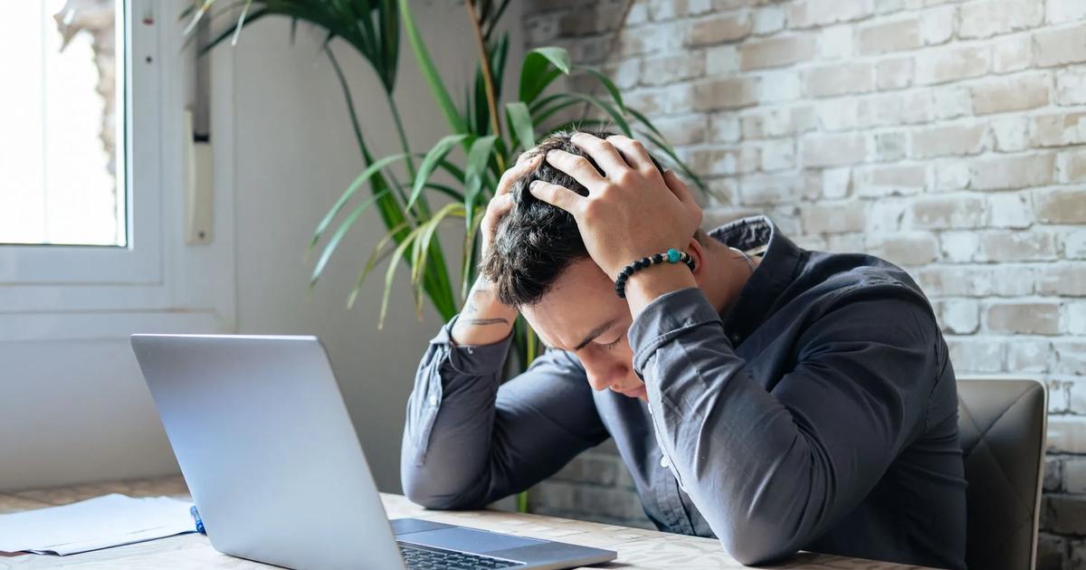 A frustrated man sits before a laptop with his hands over his head