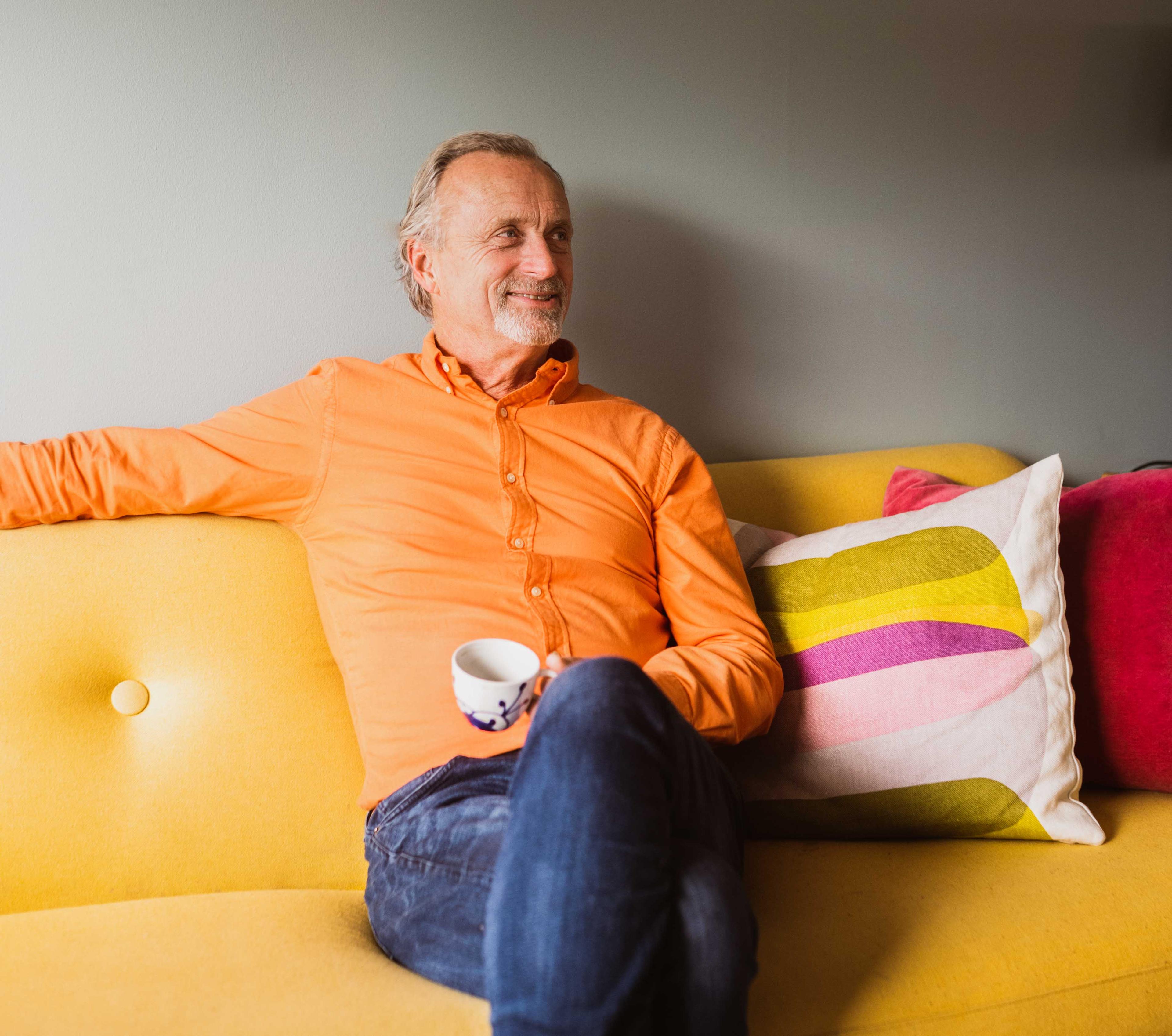 En avslappet og tilfreds middelaldrende mann med kaffekopp som sitter i en gul sofa. Han virker fornøyd med endelig å ha skrevet en fremtidsfullmakt!