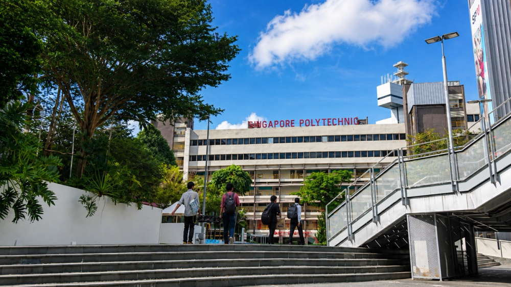 Four team members walking up an outdoor staircase toward the Singapore Polytechnic building, one carrying equipment, under a clear blue sky
