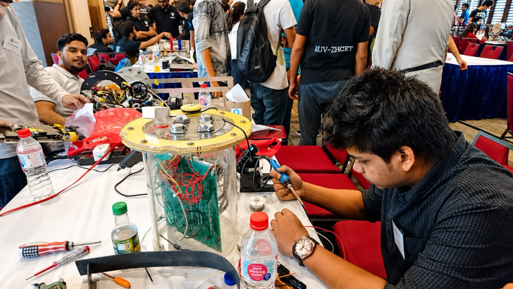 Adnan soldering a wire while sitting beside BRACU Duburi's open transparent enclosure, with other SAUVC teams working at nearby tables in a shared assembly hall