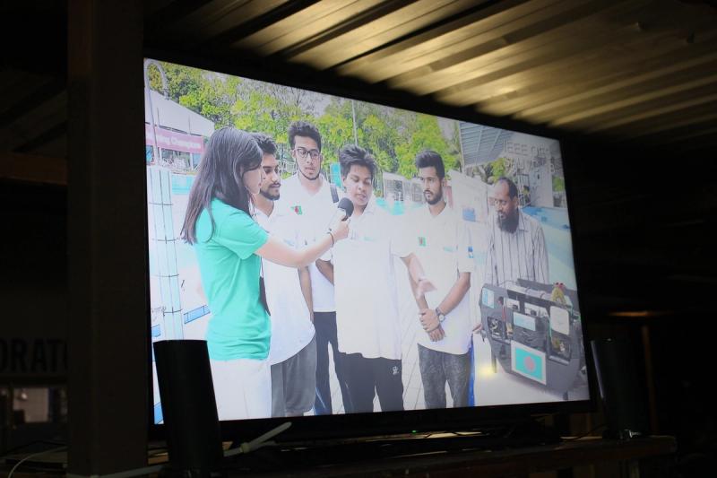 A TV screen displaying video footage of a reporter interviewing four BRACU Duburi team members and Dr. Khalilur Rahman poolside, with a SAUVC banner and the Bangladesh-flagged robot visible in the background