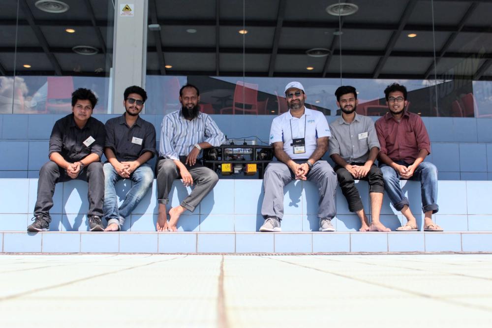 Six people sitting on the poolside edge with BRACU Duburi placed between them, including Dr. Khalilur Rahman barefoot on the left and a man in a white SAUVC polo shirt and cap in the center
