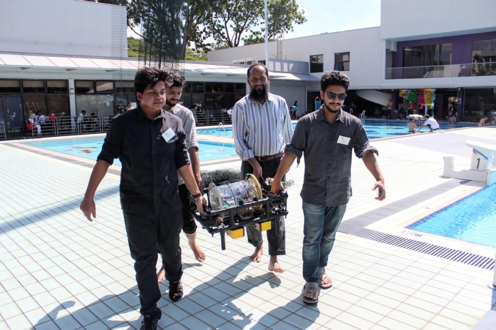 Four team members, one barefoot, carrying BRACU Duburi's transparent cylindrical enclosure past a swimming pool complex, with Dr. Khalilur Rahman in the middle of the group