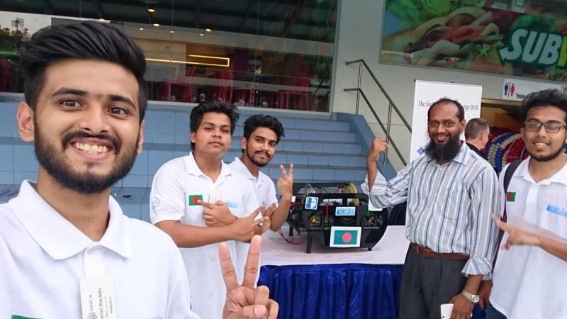 Team members in white BRACU Duburi polo shirts making victory signs with Dr. Khalilur Rahman, Duburi displayed on a table with a Bangladesh flag behind them