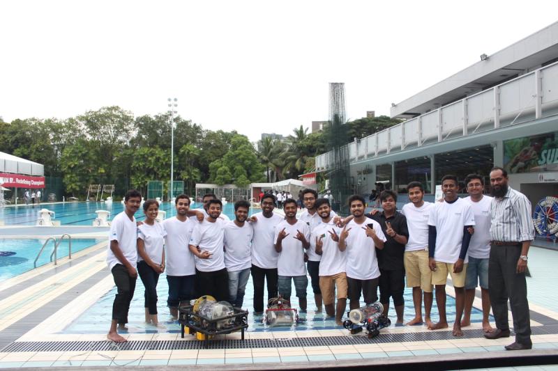 A large group of SAUVC teams in matching white shirts standing together poolside with three underwater robots placed in front of them