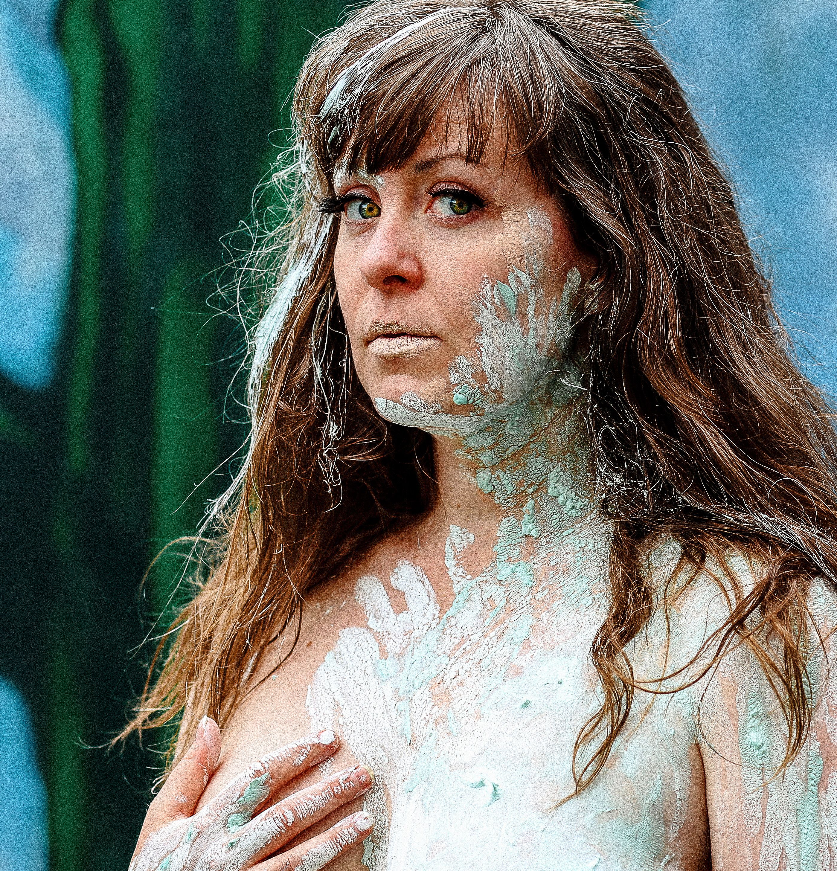 A white woman with long brown hair with grey strands looks slightly aside at the camera. They are topless with just the upper part of their chest visible, and one hand covering one breast. Light green paint is smeared across their chest, shoulder, neck, and face with some in their hair. Human handprints are discernible in the dried paint.
