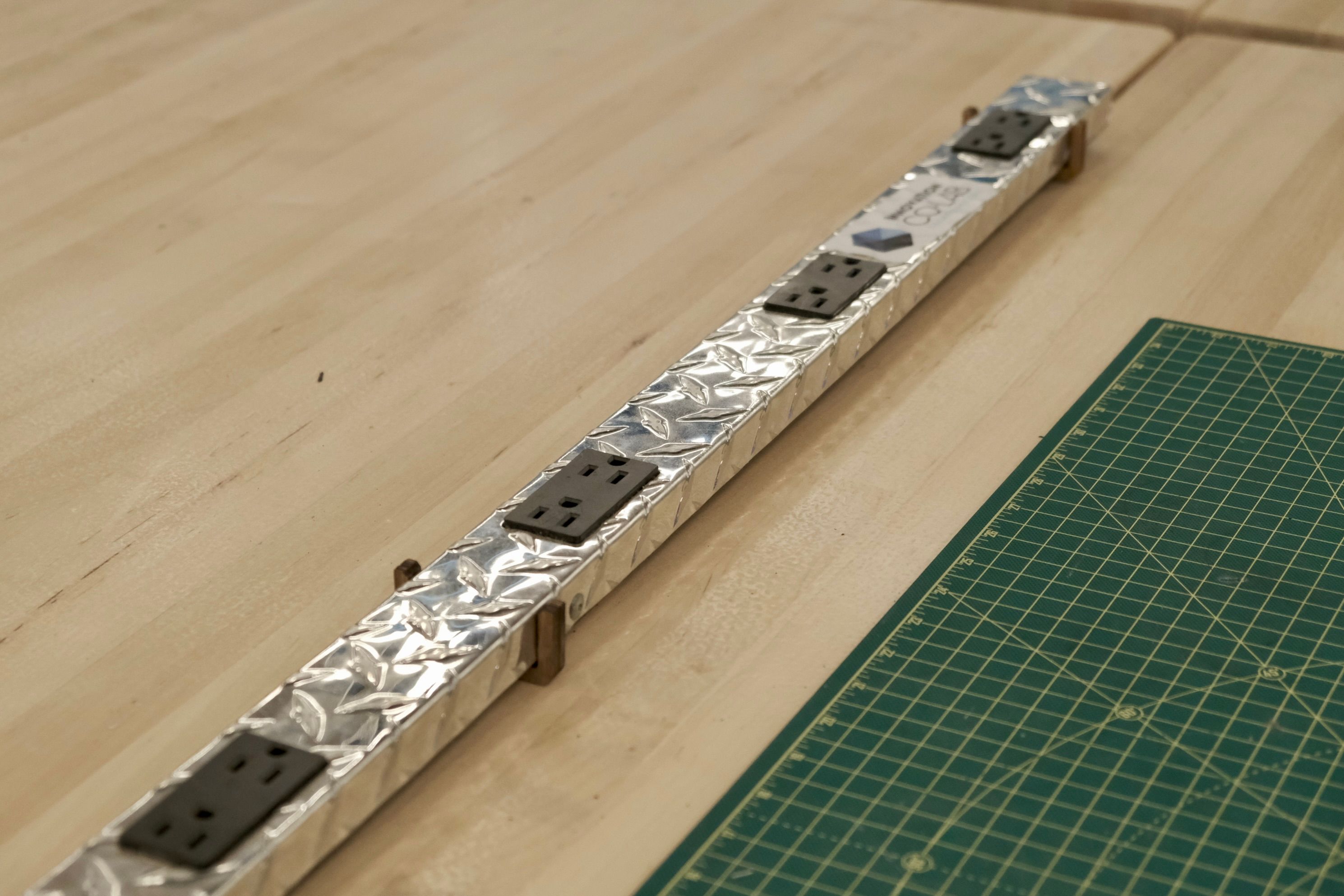 a long metal power strip with black outlets held between two wooden tables with laser cut parts