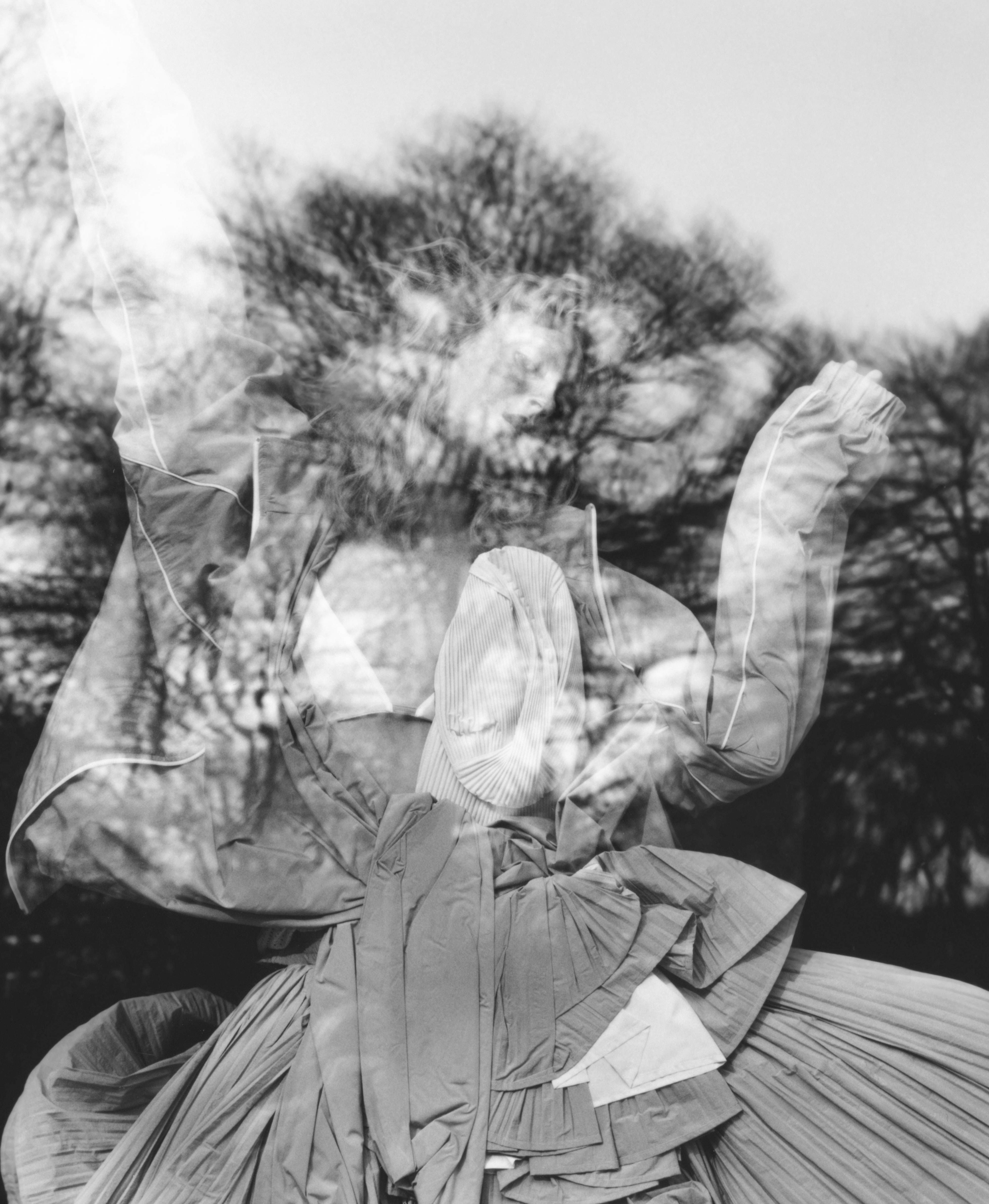 Black-and-white double exposure of a person in a pleated, sculptural dress with leafless trees overlaid, arms raised in motion.