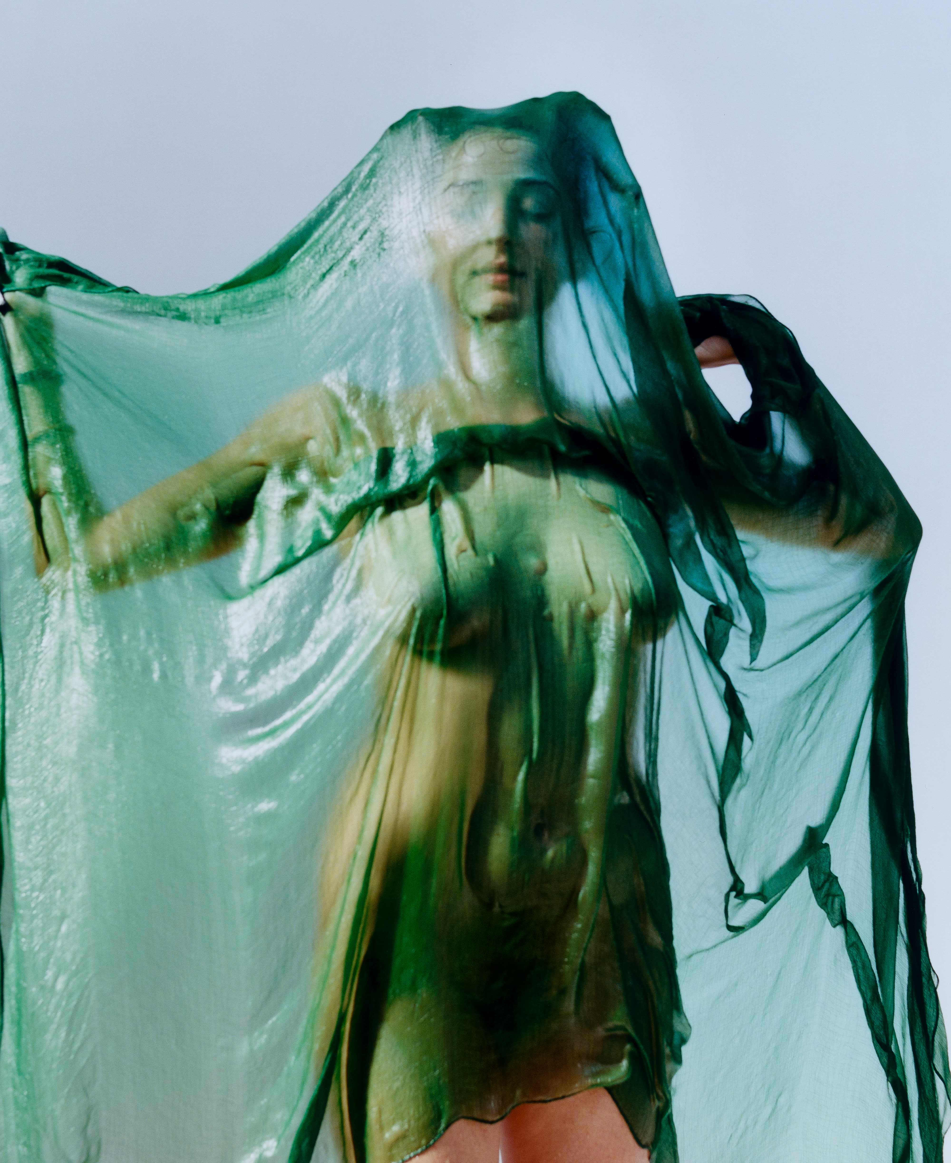 Portrait of a person draped in translucent green fabric, arms lifting the veil, face softly visible against a pale sky.