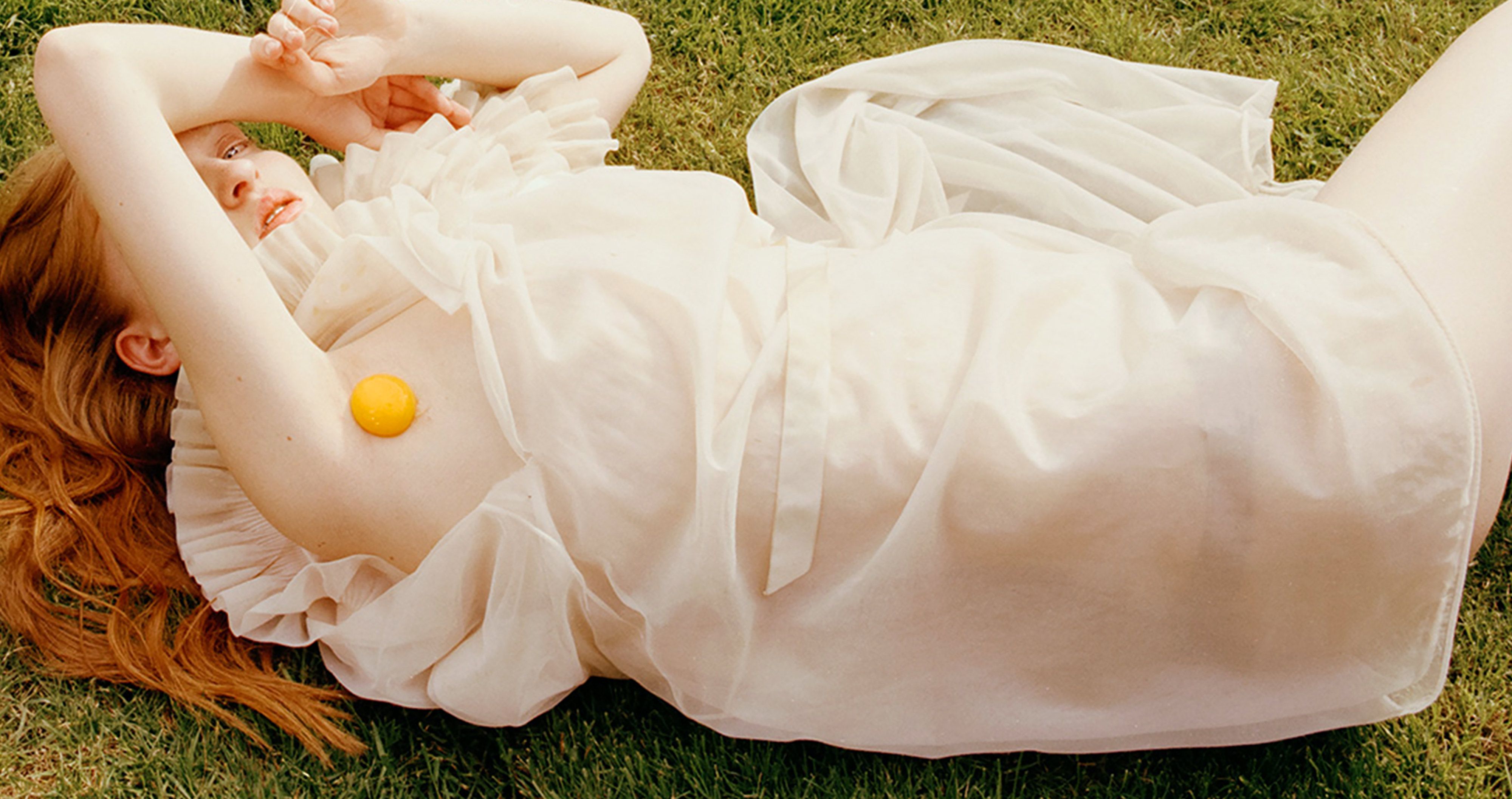 Sunlit portrait of a red-haired person lying on grass in a sheer white dress, a small lemon tucked under the arm, arms raised overhead.