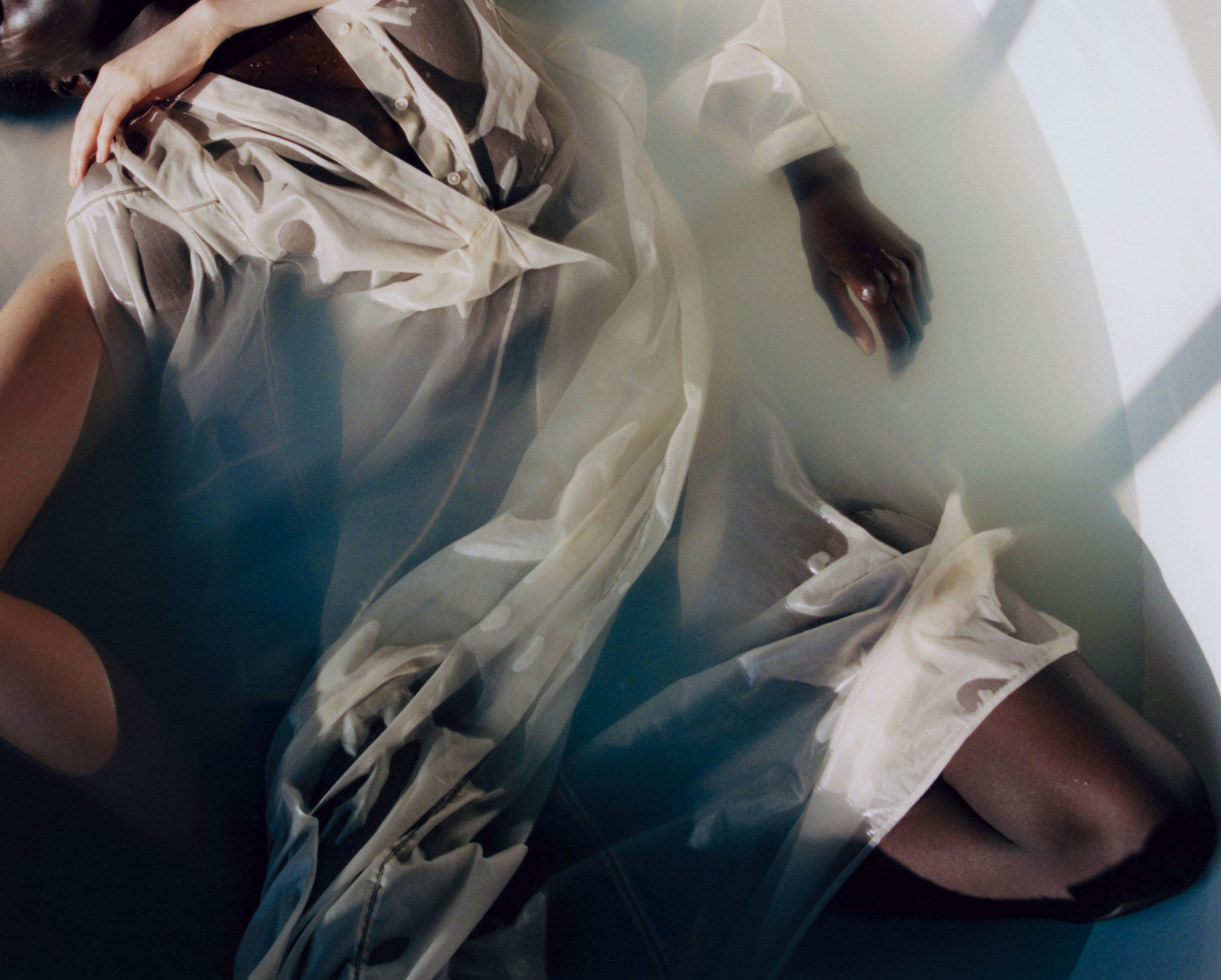 Overhead photo of a figure reclining in milky water, wearing a sheer white dress, soft light and shadows pooling across the fabric.
