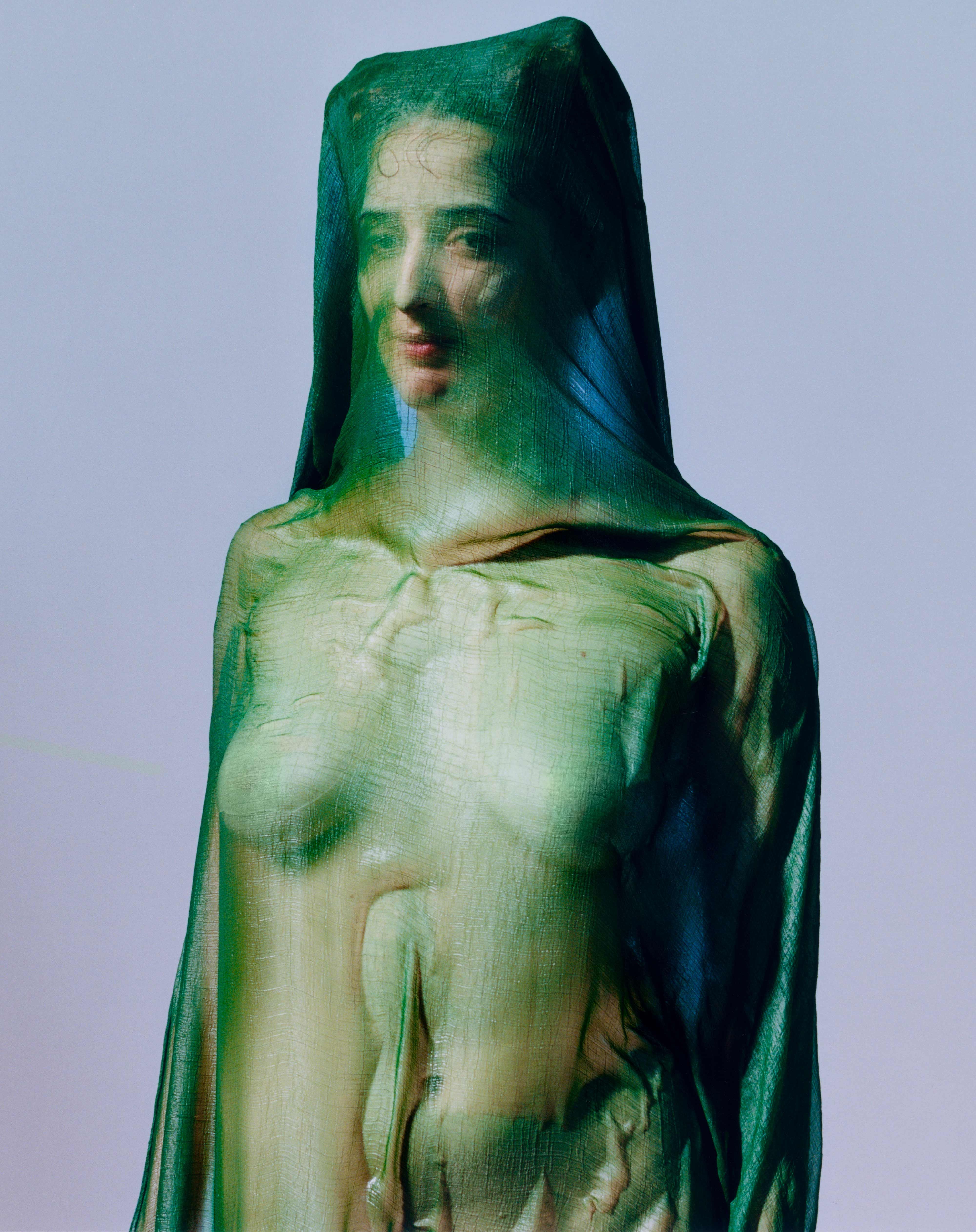 Portrait of a person draped head-to-toe in translucent green fabric, features softly visible, against a pale background.
