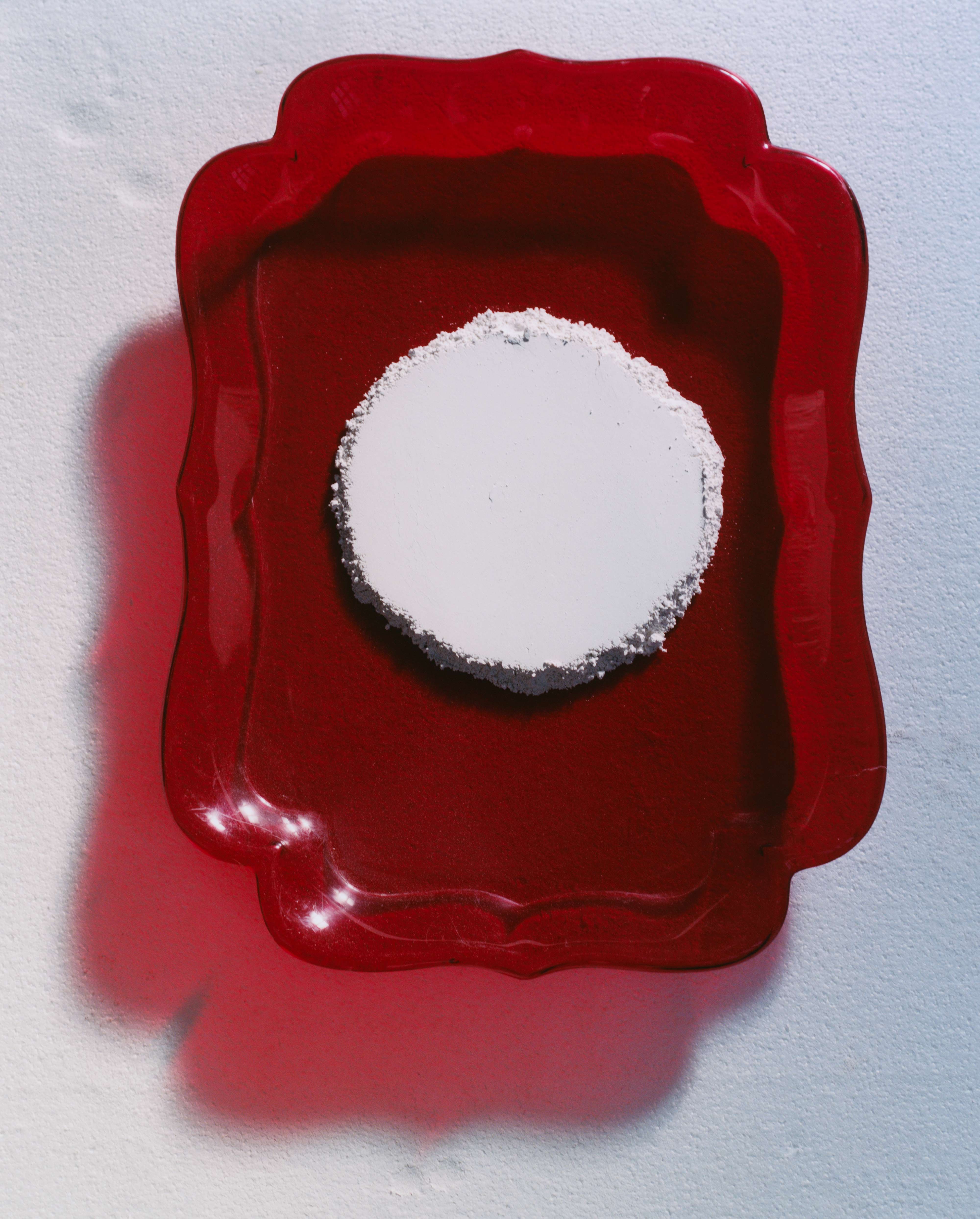 Top-down still life: scalloped red tray holding a white circular powder puck on a powder-dusted surface with soft shadow.