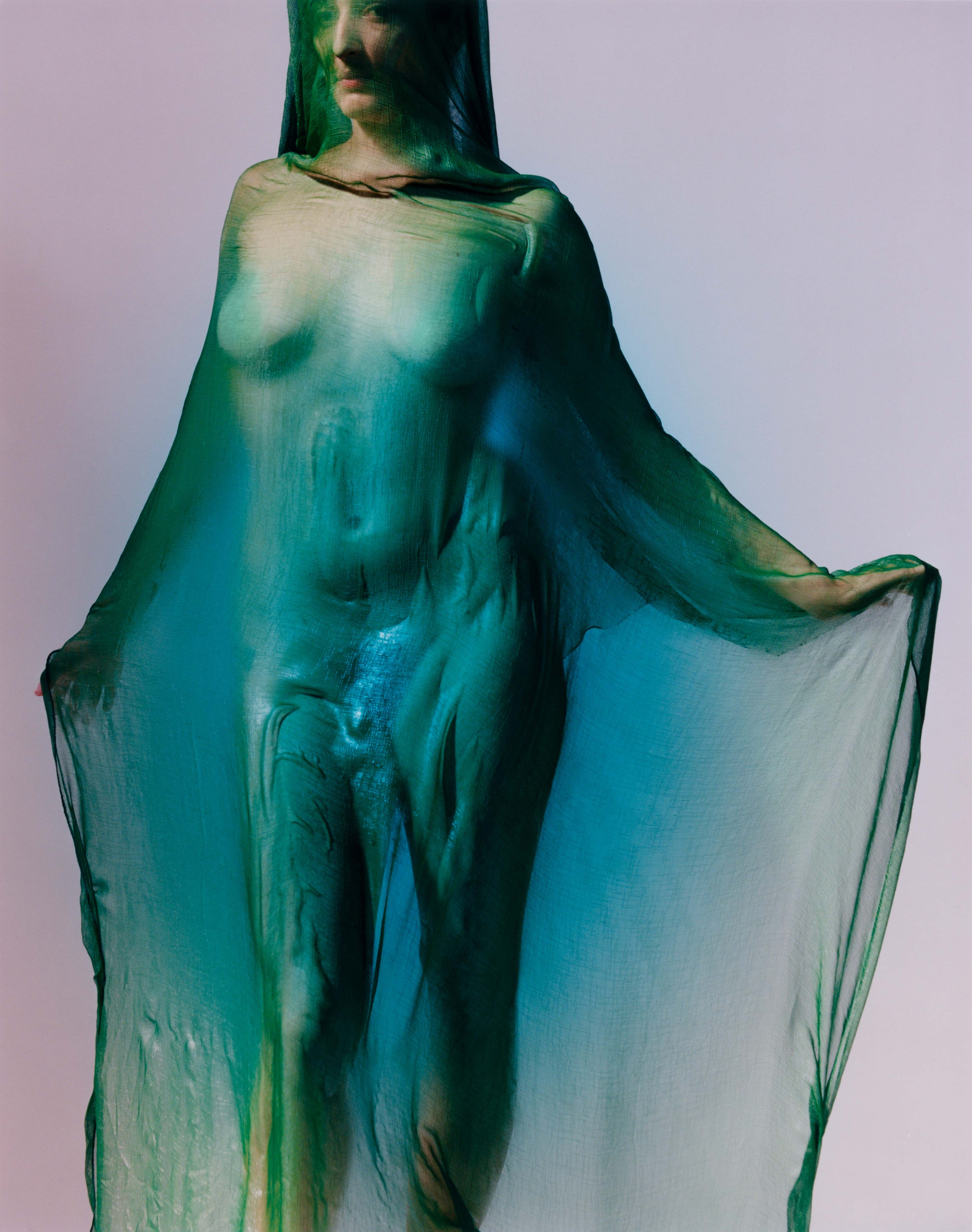 Full-length portrait of a person draped in translucent green fabric, holding the veil out to the sides, features softly visible against a pale backdrop.