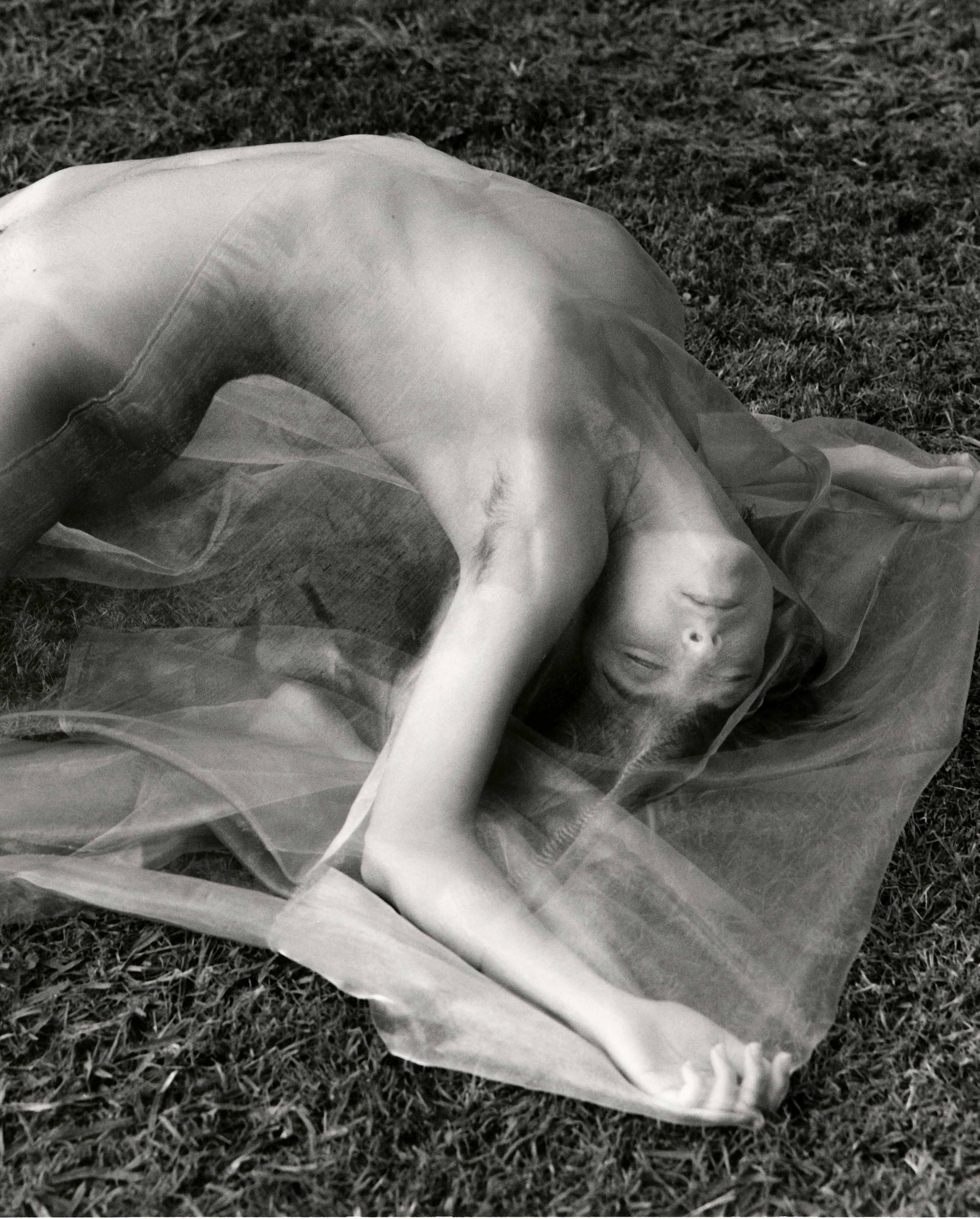 Black-and-white photo of a person lying on grass beneath a sheer veil, back arched with arms extended, face softly obscured.