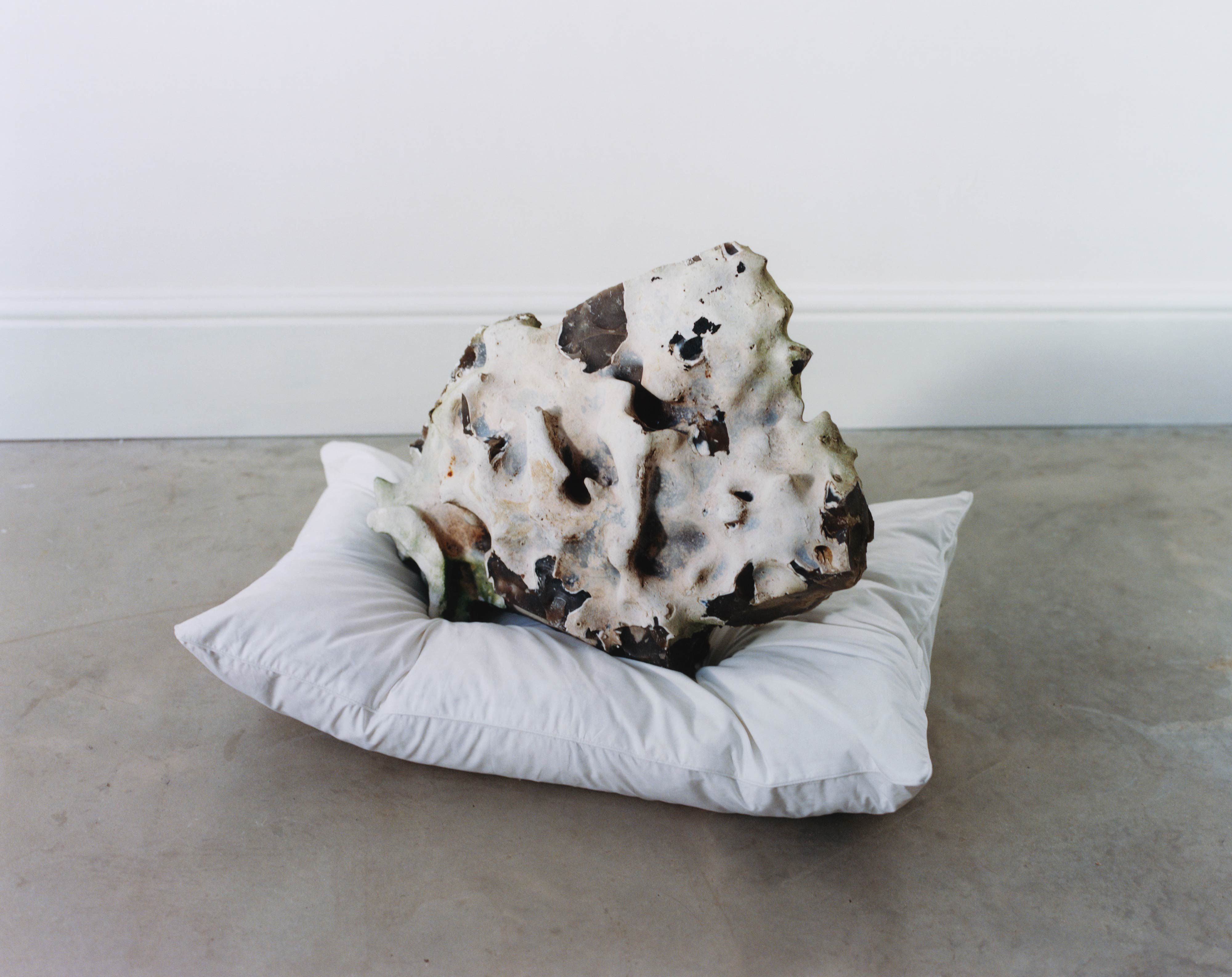 Porous, sculptural rock resting on a white pillow on a concrete floor against a plain wall.