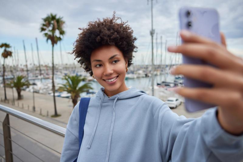 young-curly-haired-teenage-girl-makes-selfie-modern-smartphone-smiles-pleasantly-wears-hoodie-carries-rucksack-poses-against-harbor-stands-pier-spends-free-time-embankment.jpg