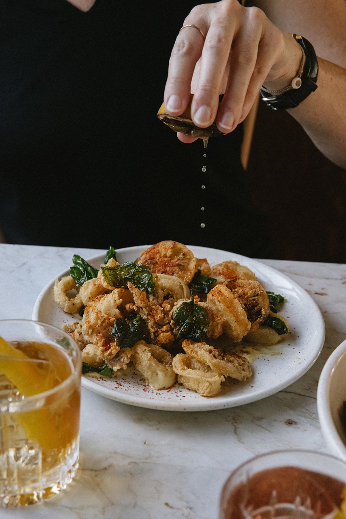 A hand squeezes lemon over a plate of fried calamari with crispy sage, beside amber cocktails on a marble table