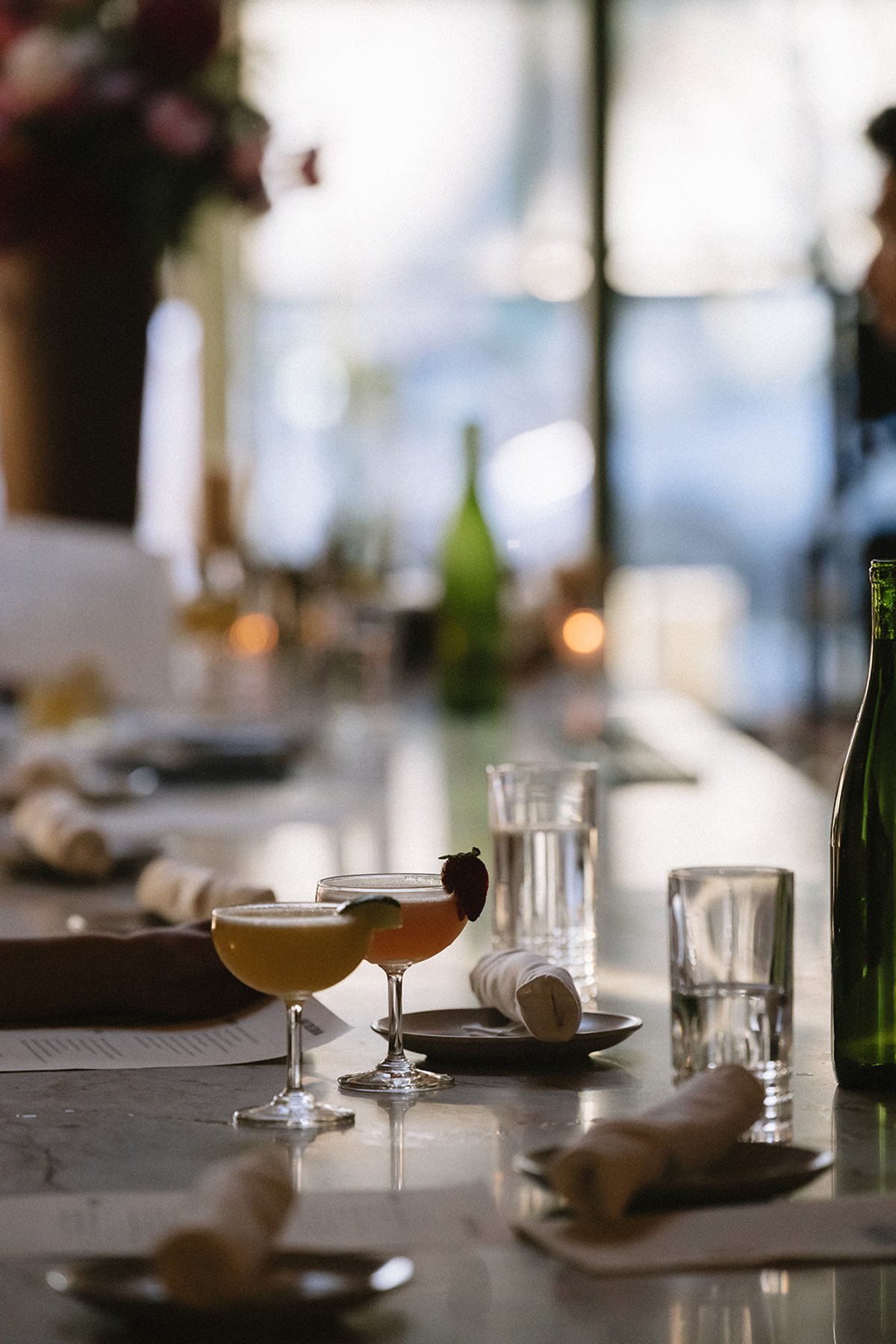 Two garnished coupe cocktails on a marble bar, with water glasses and a green wine bottle softly blurred behind them