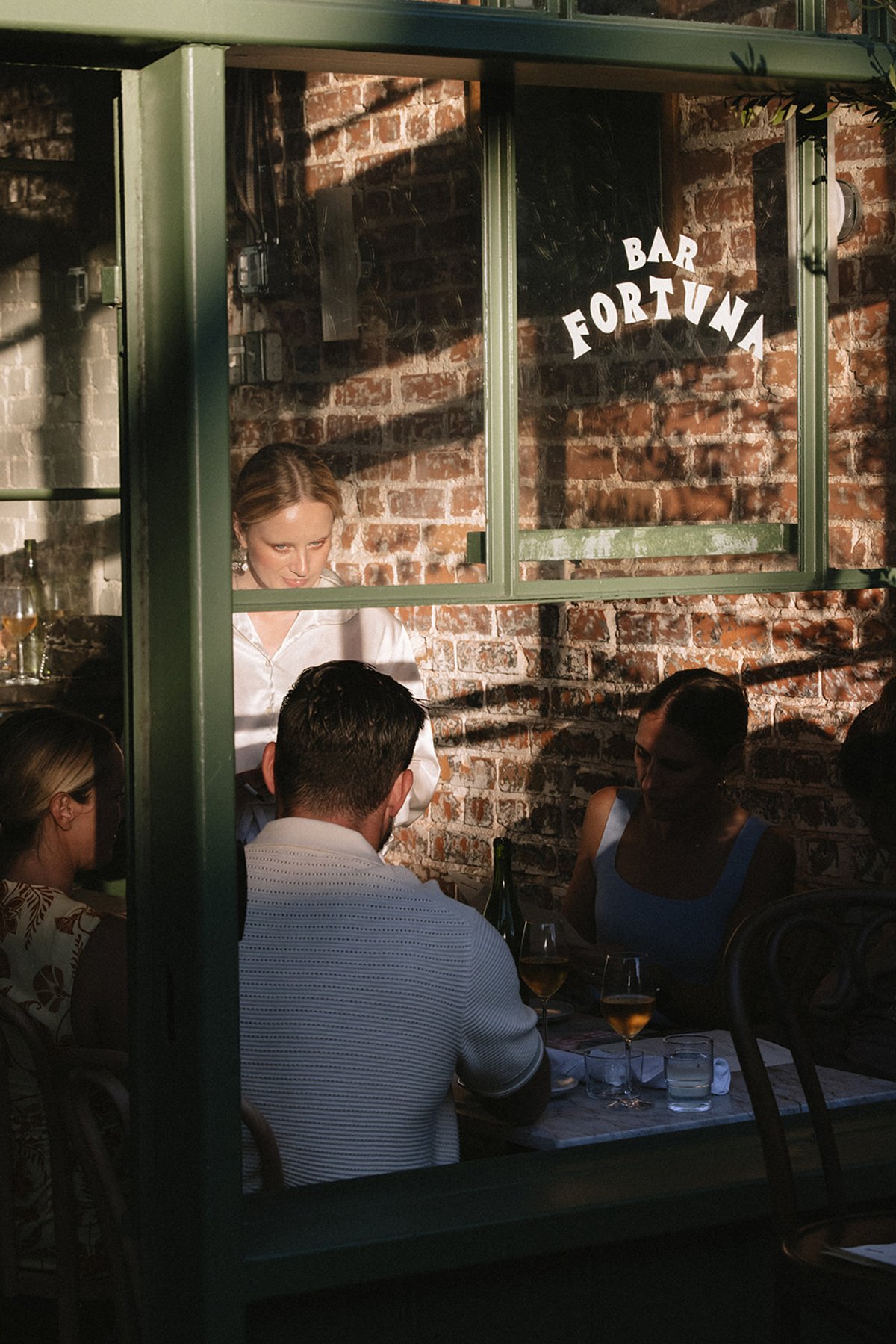 Bar Fortuna's gold window lettering, with a server greeting guests seated at a table inside