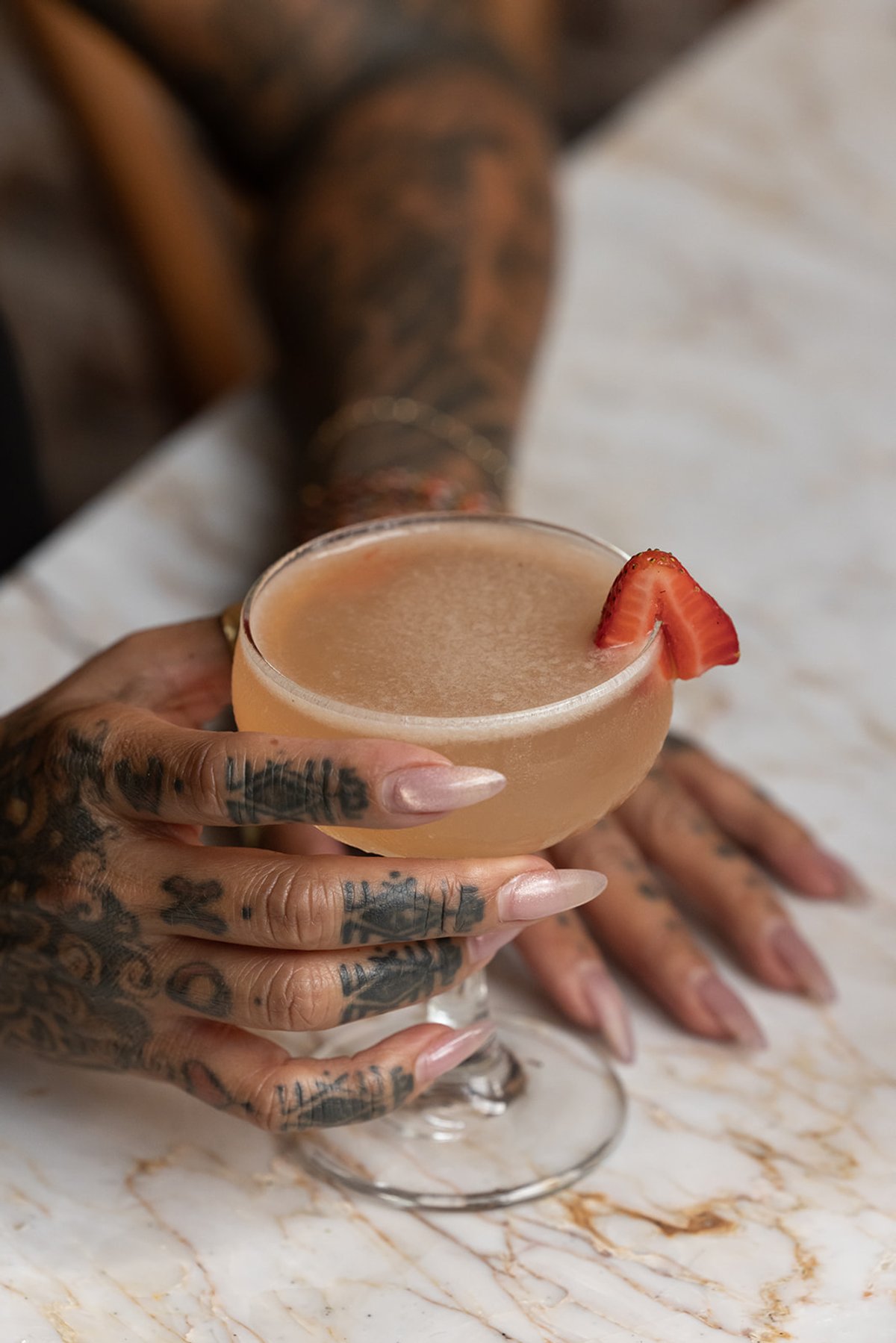 A tattooed hand holds a strawberry-garnished cocktail in a coupe glass on a marble table