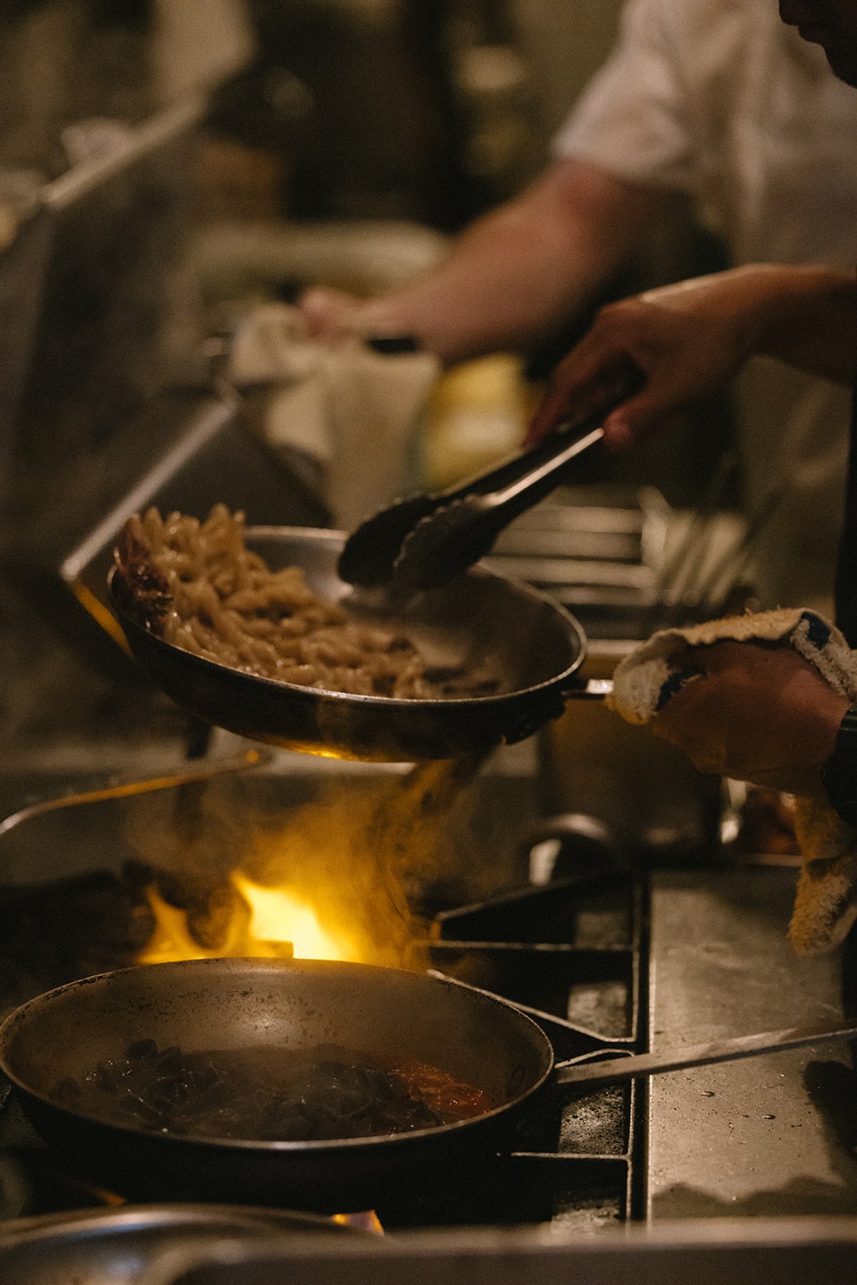 A cook tosses pasta in a flaming pan over an open flame on the kitchen line