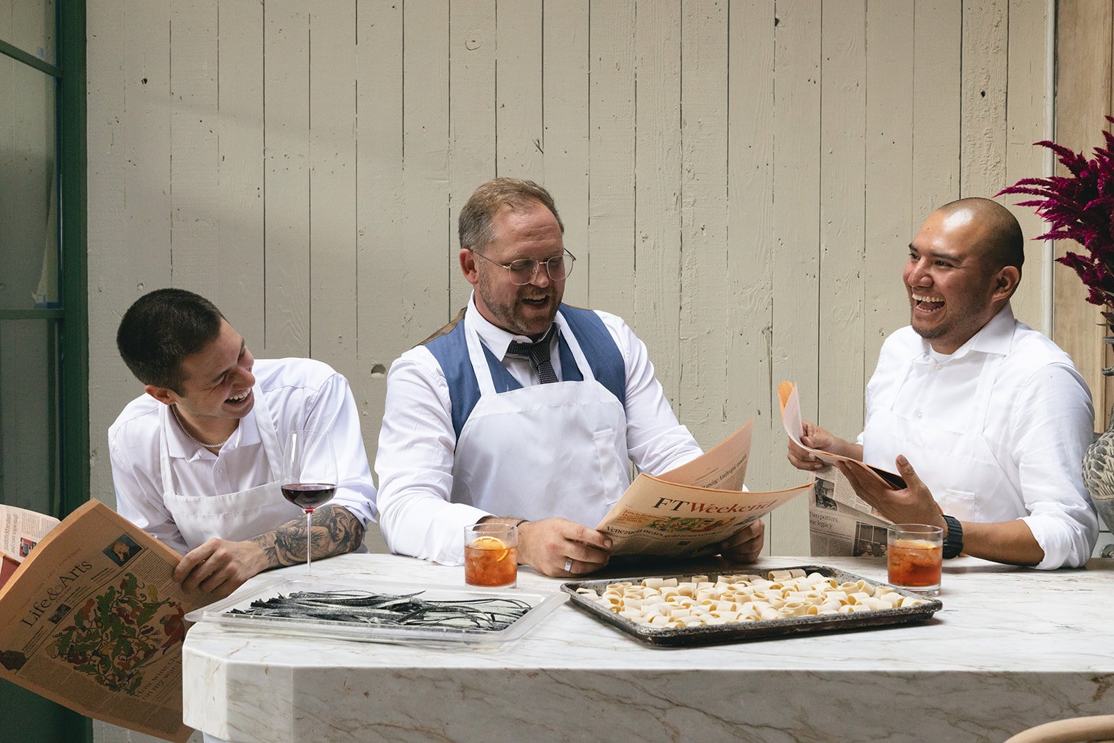 Three Bar Fortuna team members laughing together at the marble bar, reading newspapers over trays of fresh pasta and cocktails