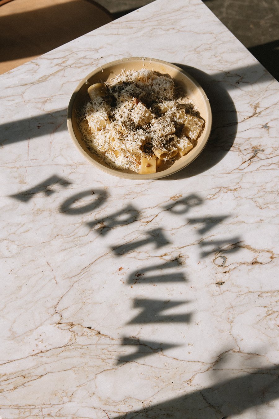 A plate of pasta topped with shaved cheese and truffle, the shadow of Bar Fortuna's window lettering cast across a sunlit marble table