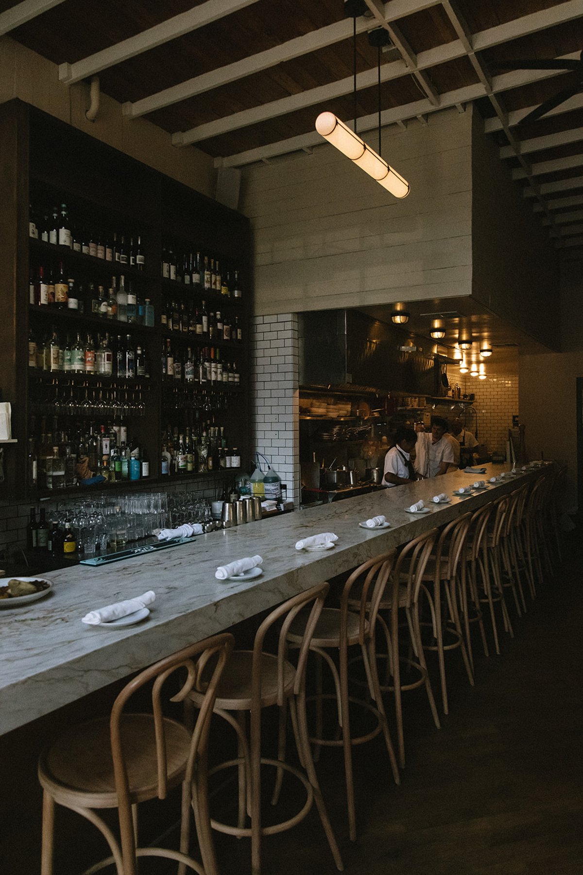 Bar Fortuna's marble bar with bentwood stools, bottle-lined shelves, and the open kitchen glowing beyond