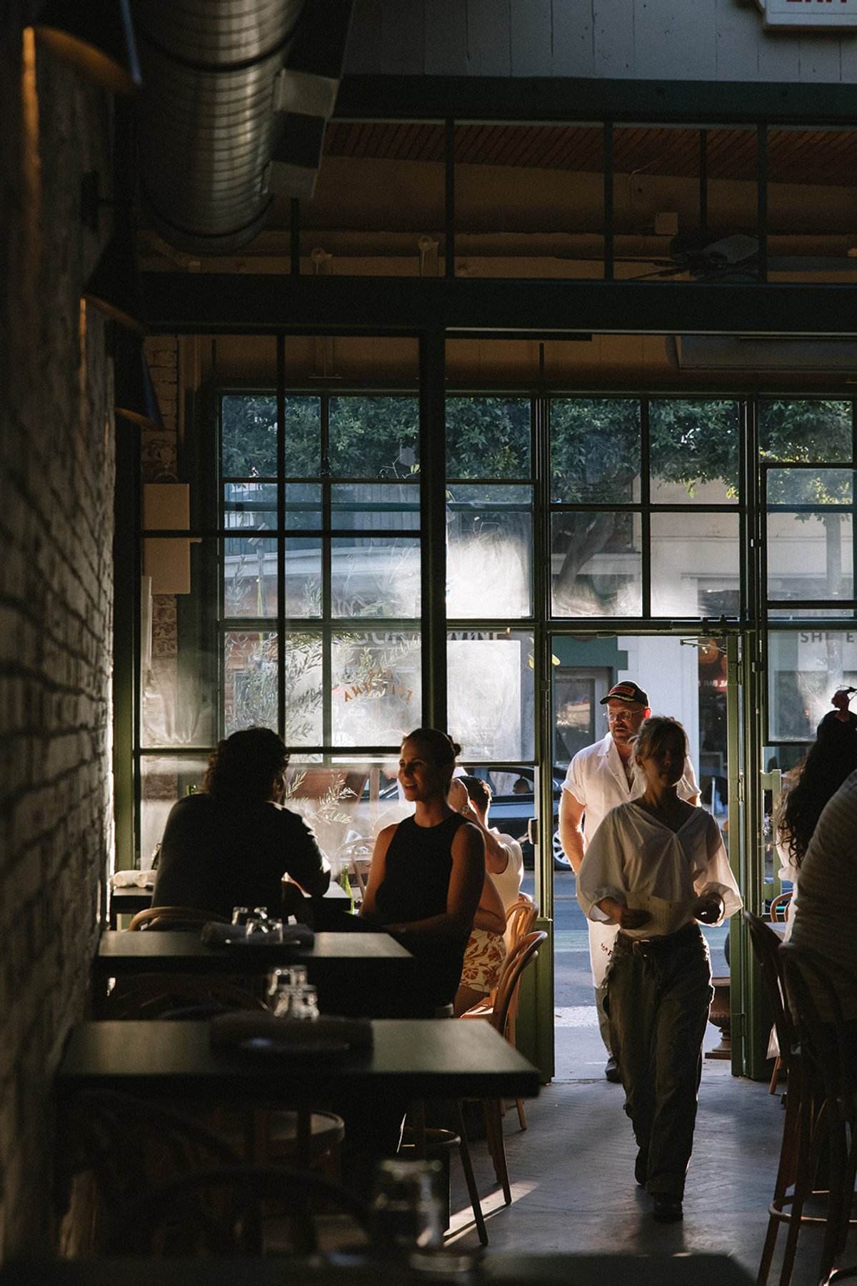 Guests seated at tables and walking in through Bar Fortuna's open storefront doors at golden hour, trees visible through the windows