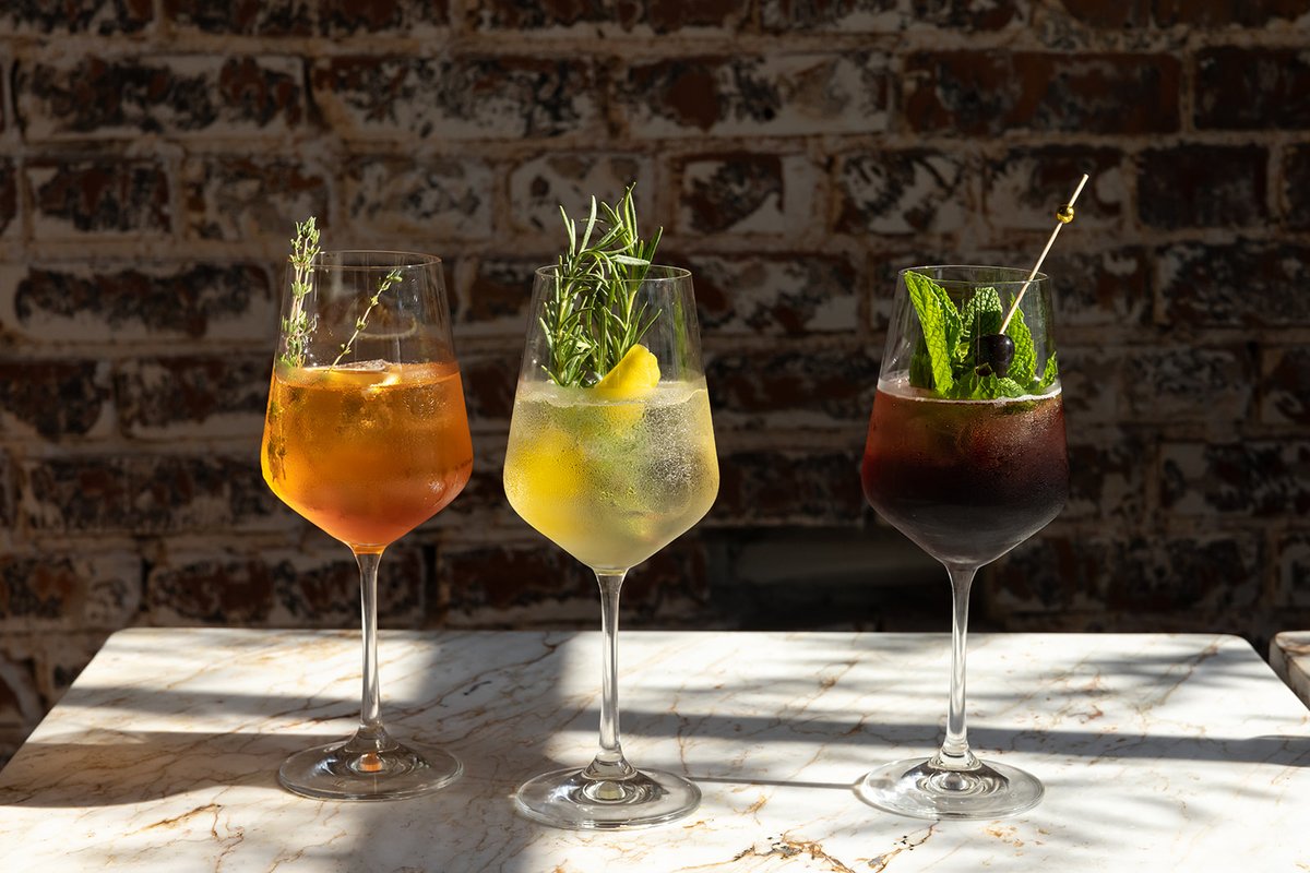 Three cocktails lined up on a marble table against an exposed brick wall — an amber spritz with thyme, a pale herb-garnished spritz, and a dark red cocktail with mint and a cherry