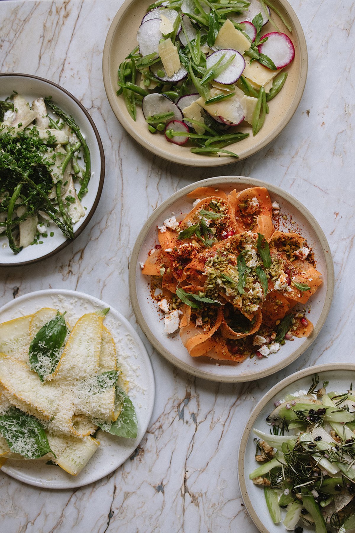 An overhead view of five composed salads on marble, including shaved carrot with pistachio and feta, squash blossom with parmesan and sage, and snap pea with radish and shaved cheese