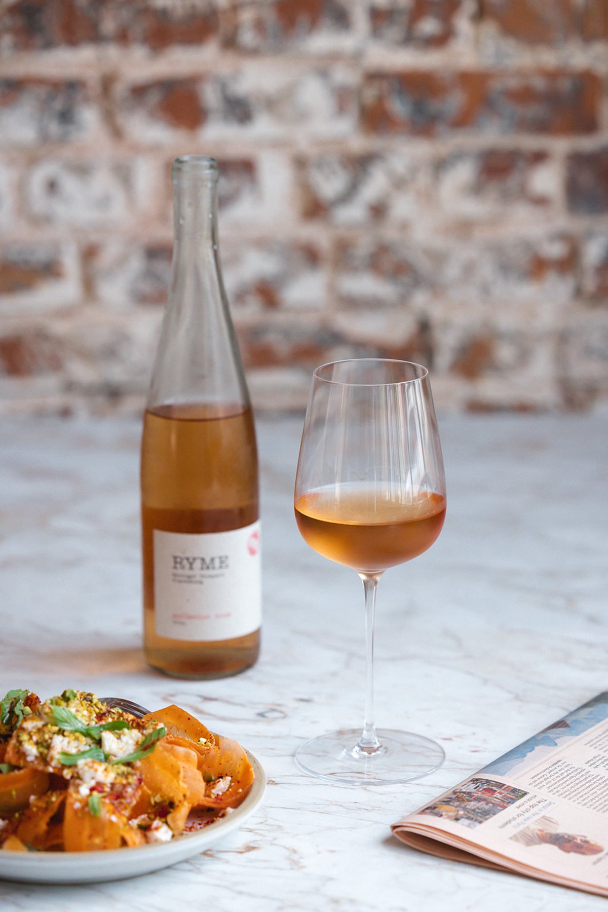 A glass and bottle of orange wine beside a plate of spiced carrot ribbons on a marble table, with a magazine open nearby