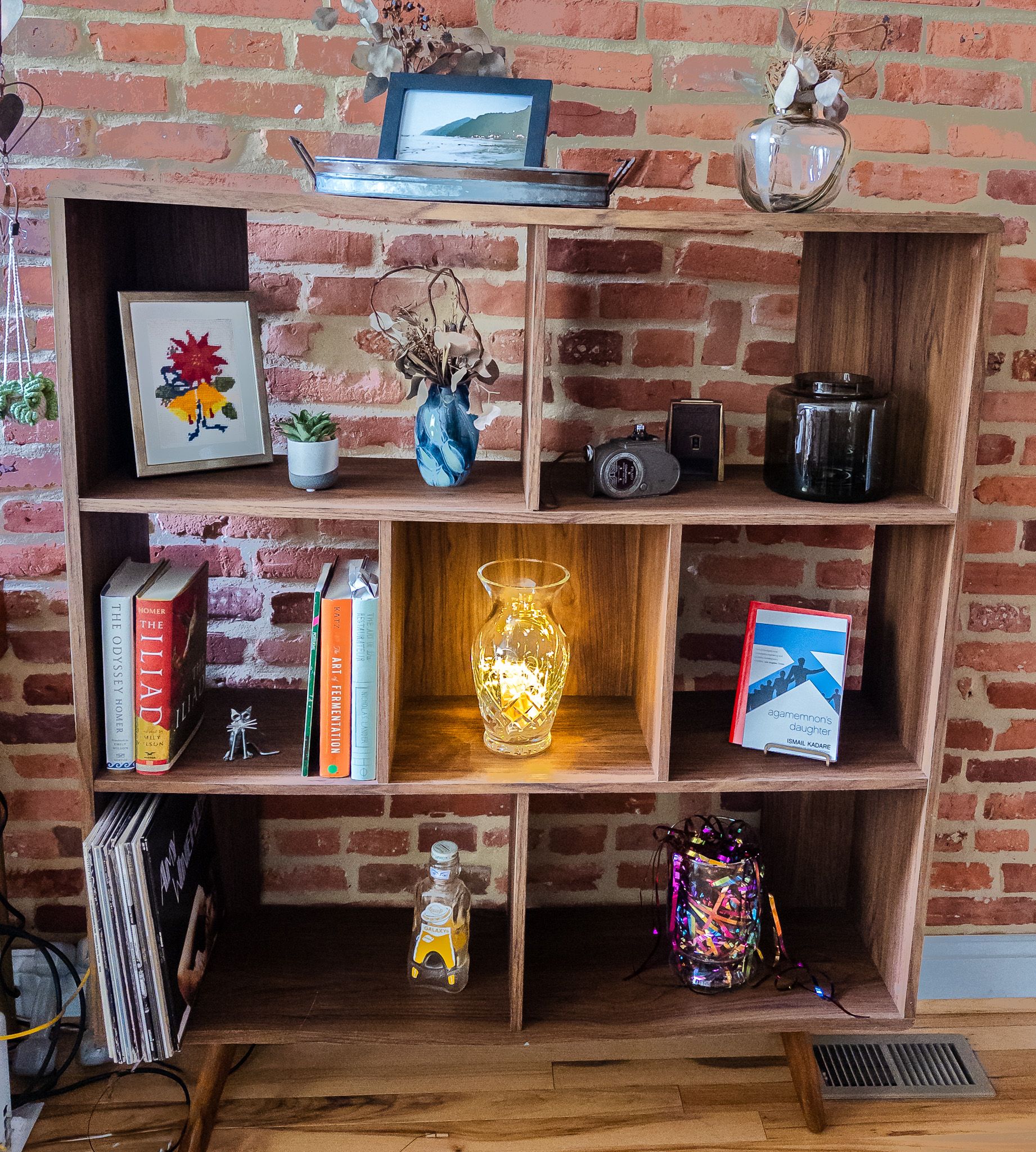 Midcentury modern walnut-toned bookshelf with staggered open cubbies against an exposed red brick wall, styled with hardcover books, framed art, dried-flower vases, a glowing lamp, and vinyl records.