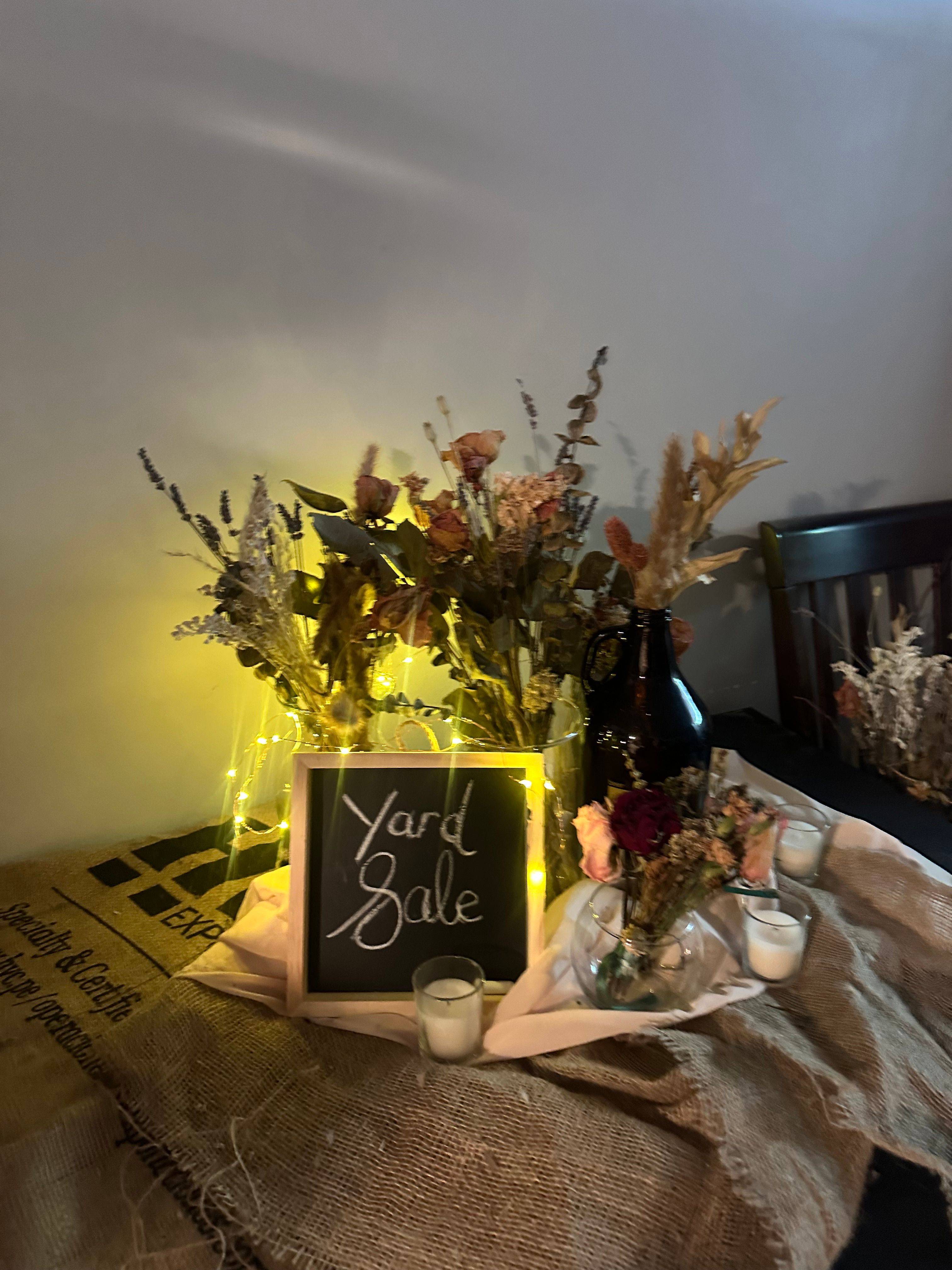 Dried-flower arrangements in glass vases and a dark glass jug, glowing with fairy lights beside a "Yard Sale" chalkboard sign.