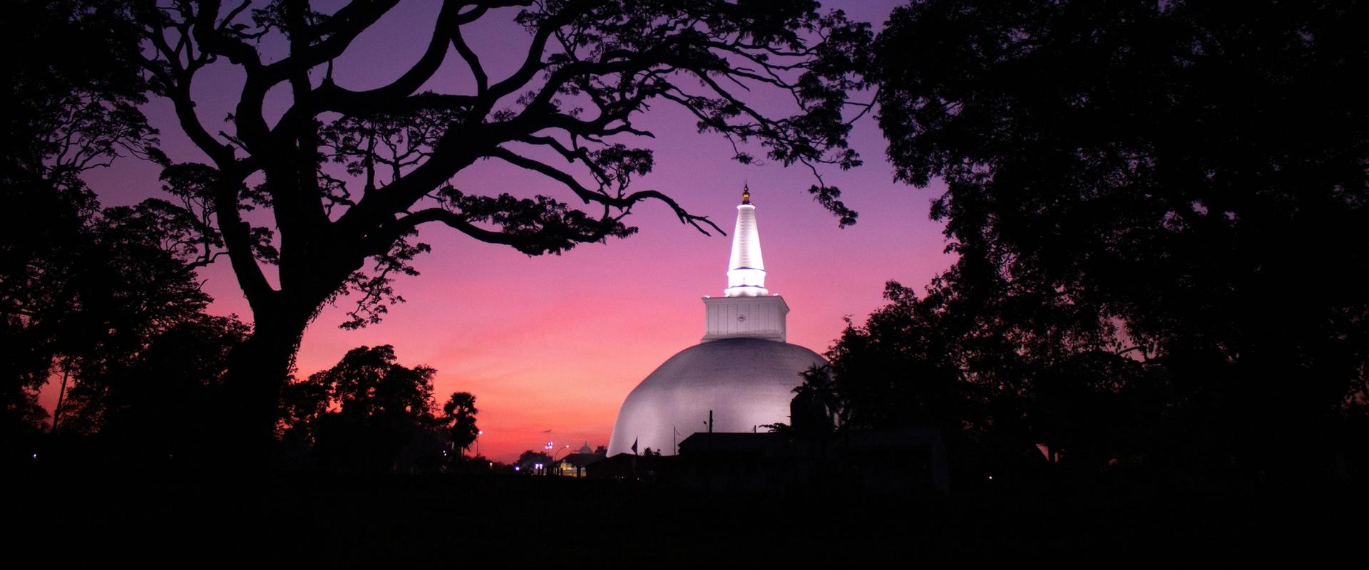 Anuradhapura Ruwanwelisaya stupa at sunset, Sri Lanka – white Buddhist dagoba illuminated against a pink and purple twilight sky framed by dark tree silhouettes in the Sacred City UNESCO site.