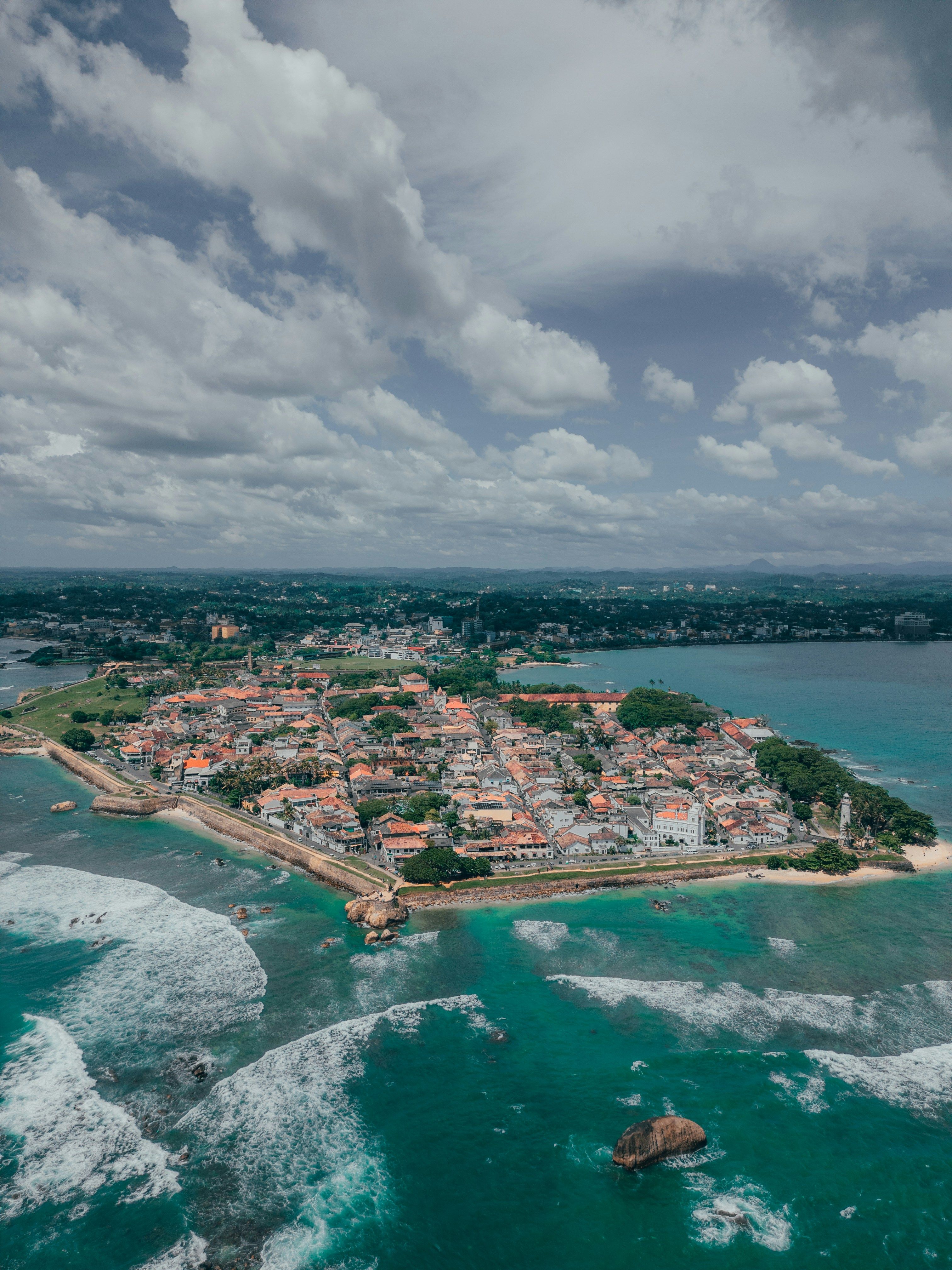 Drone aerial view of the historic Galle Fort peninsula showing colonial-era buildings with red rooftops, fortified walls, turquoise ocean waves, and the Galle city skyline in the background, southern Sri Lanka