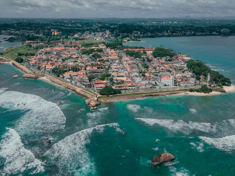 Drone aerial view of the historic Galle Fort peninsula showing colonial-era buildings with red rooftops, fortified walls, turquoise ocean waves, and the Galle city skyline in the background, southern Sri Lanka