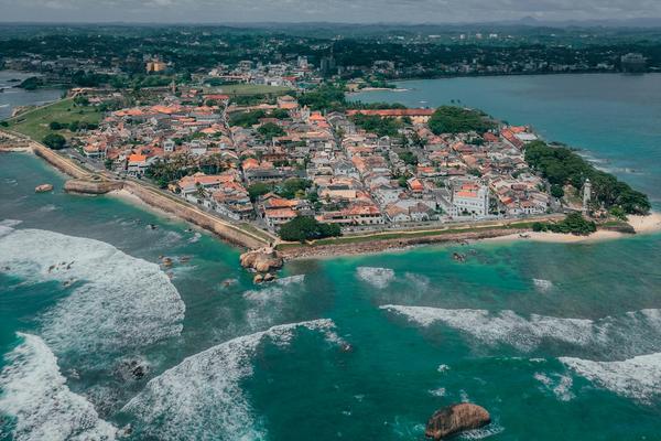Drone aerial view of the historic Galle Fort peninsula showing colonial-era buildings with red rooftops, fortified walls, turquoise ocean waves, and the Galle city skyline in the background, southern Sri Lanka