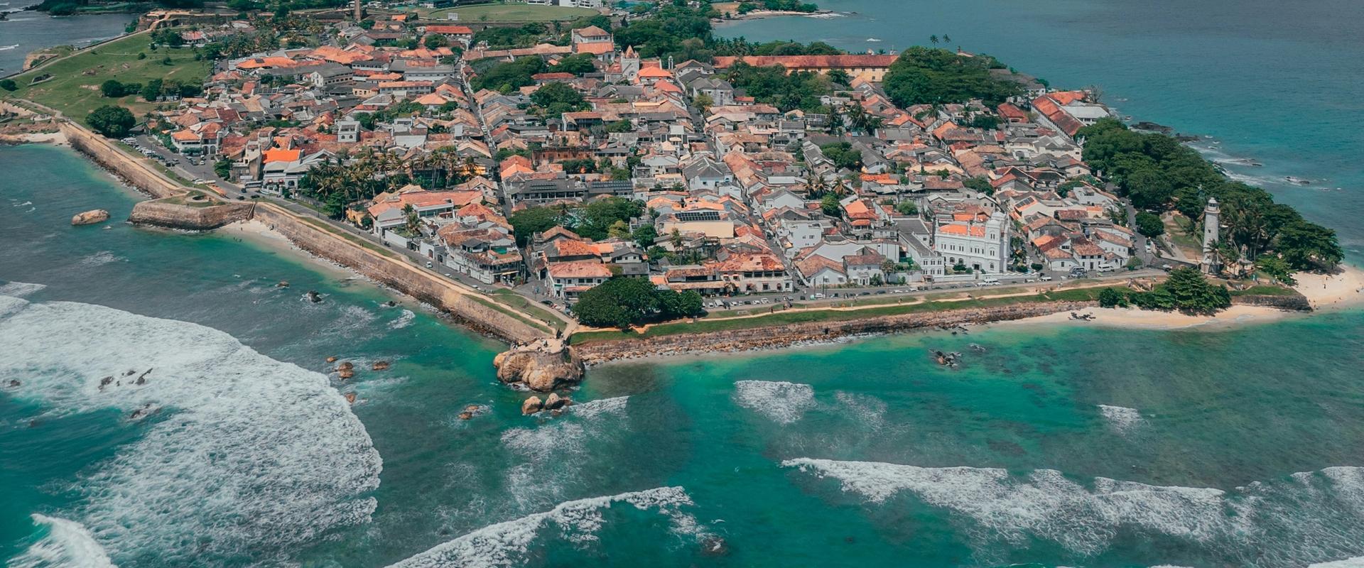 Drone aerial view of the historic Galle Fort peninsula showing colonial-era buildings with red rooftops, fortified walls, turquoise ocean waves, and the Galle city skyline in the background, southern Sri Lanka