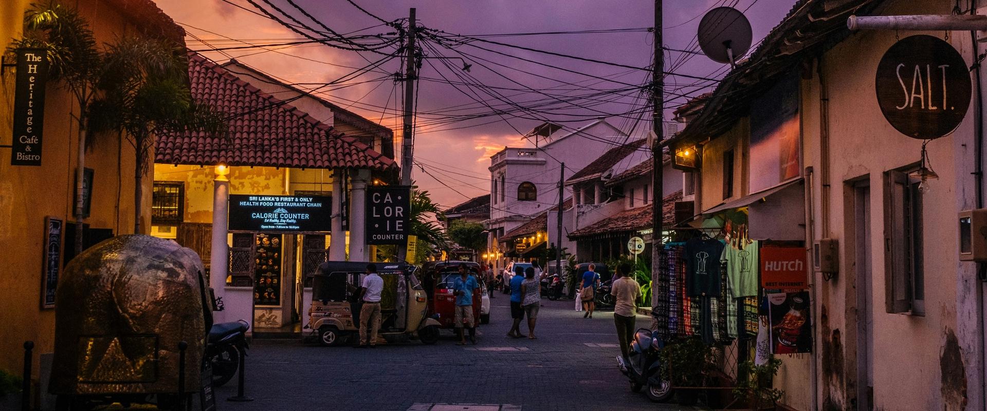 Sunset street scene inside Galle Fort, Sri Lanka, featuring colonial Dutch architecture, colorful evening sky, local shops, tuk-tuks, and tourists walking along cobblestone roads.