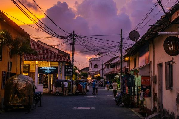 Sunset street scene inside Galle Fort, Sri Lanka, featuring colonial Dutch architecture, colorful evening sky, local shops, tuk-tuks, and tourists walking along cobblestone roads.