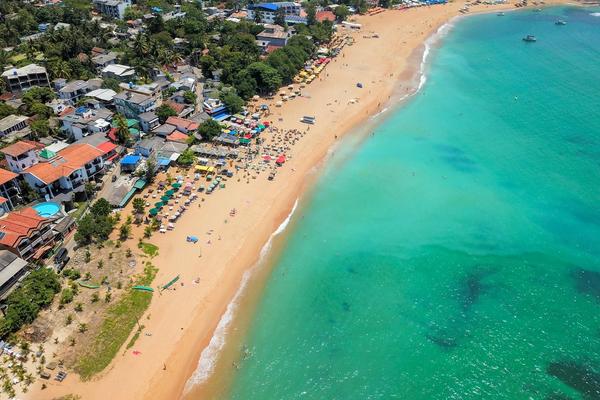 Aerial drone view of Unawatuna Beach packed with colourful sun umbrellas and swimmers in turquoise water, with the town's guesthouses and palm trees lining the golden sand, Southern Sri Lanka