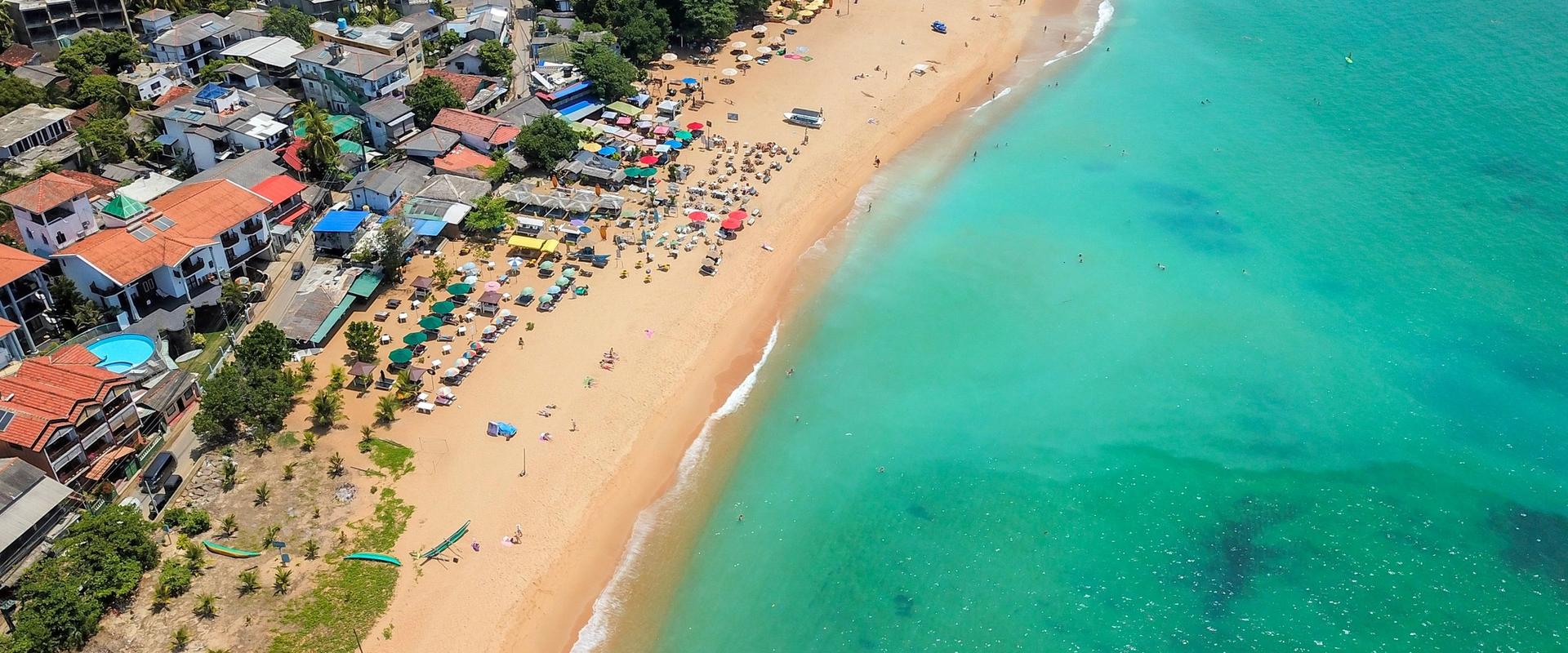 Aerial drone view of Unawatuna Beach packed with colourful sun umbrellas and swimmers in turquoise water, with the town's guesthouses and palm trees lining the golden sand, Southern Sri Lanka